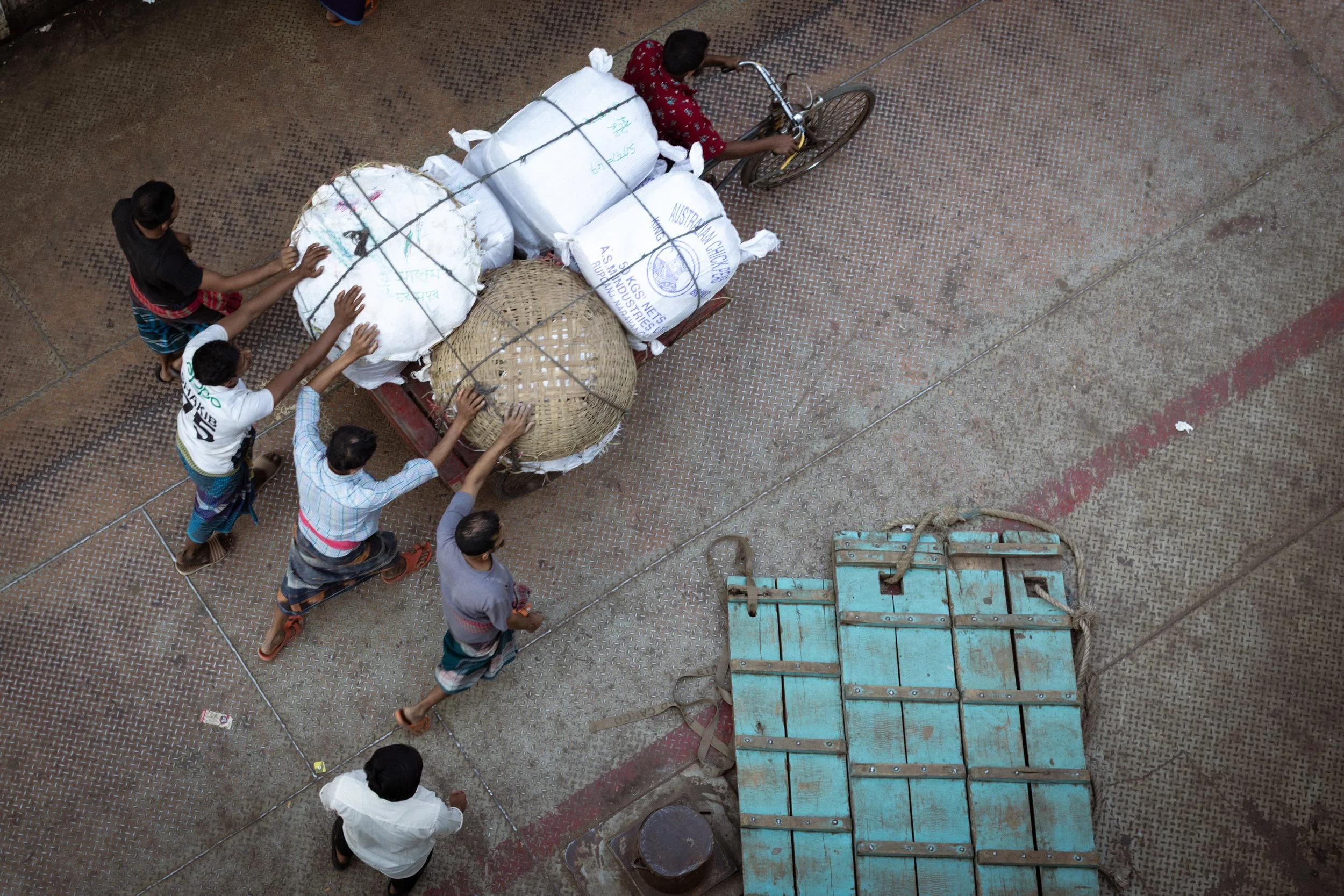Children pushing a cart loaded with large bags and woven baskets, and another cart with a blue wooden door, on a textured concrete surface.