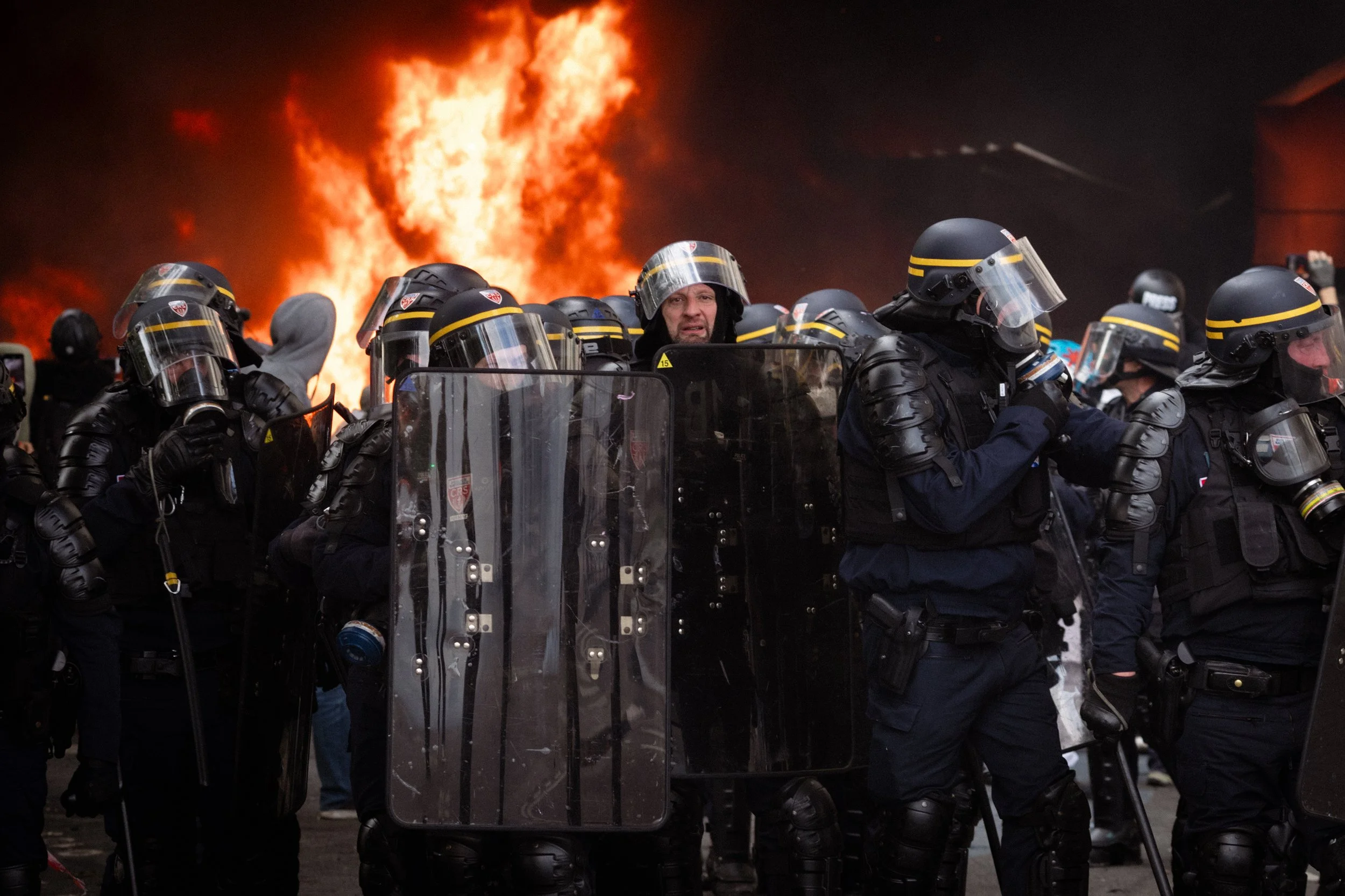 A group of riot police officers in tactical gear including helmets, shields, and protective armor, standing in front of a fiery blaze during a protest or riot.