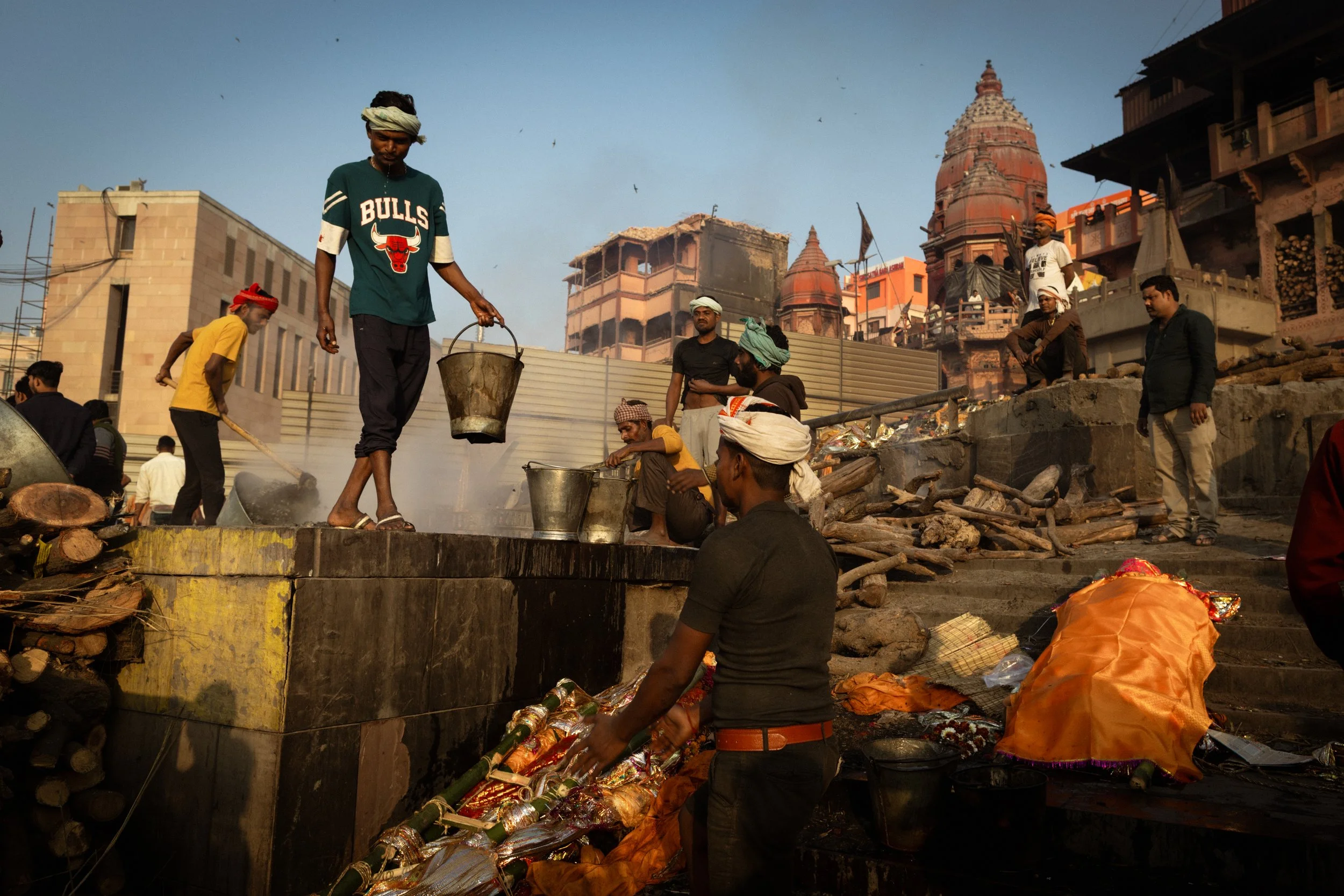 People performing a cremation ceremony at a ghat, with firewood, ritual items, and a temple in the background under a clear sky.