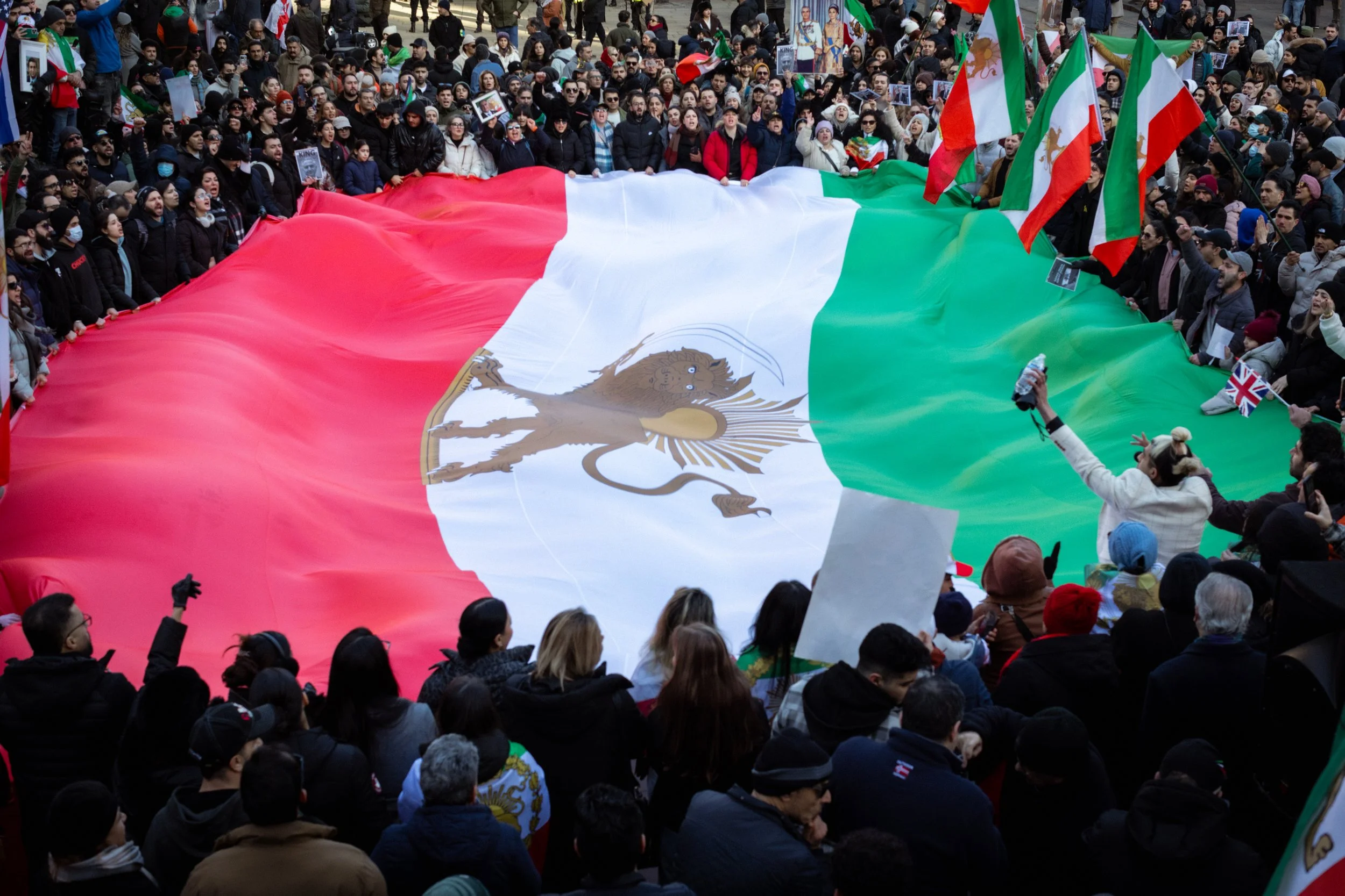 A large crowd of people gathered around a giant Mexican flag with the national emblem, the eagle holding a snake in its beak and talon, displayed in the center. Some individuals are holding and waving smaller flags.