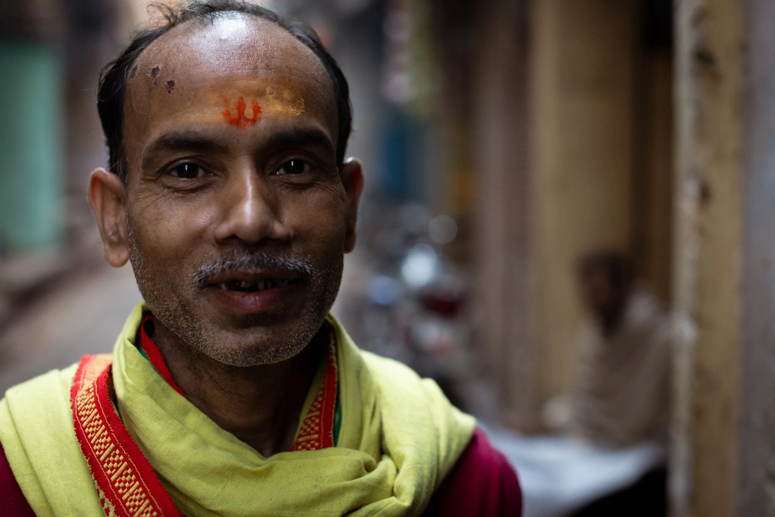 A middle-aged man with a painted red symbol on his forehead smiles, wearing a yellow and red scarf, standing outdoors in an alleyway.