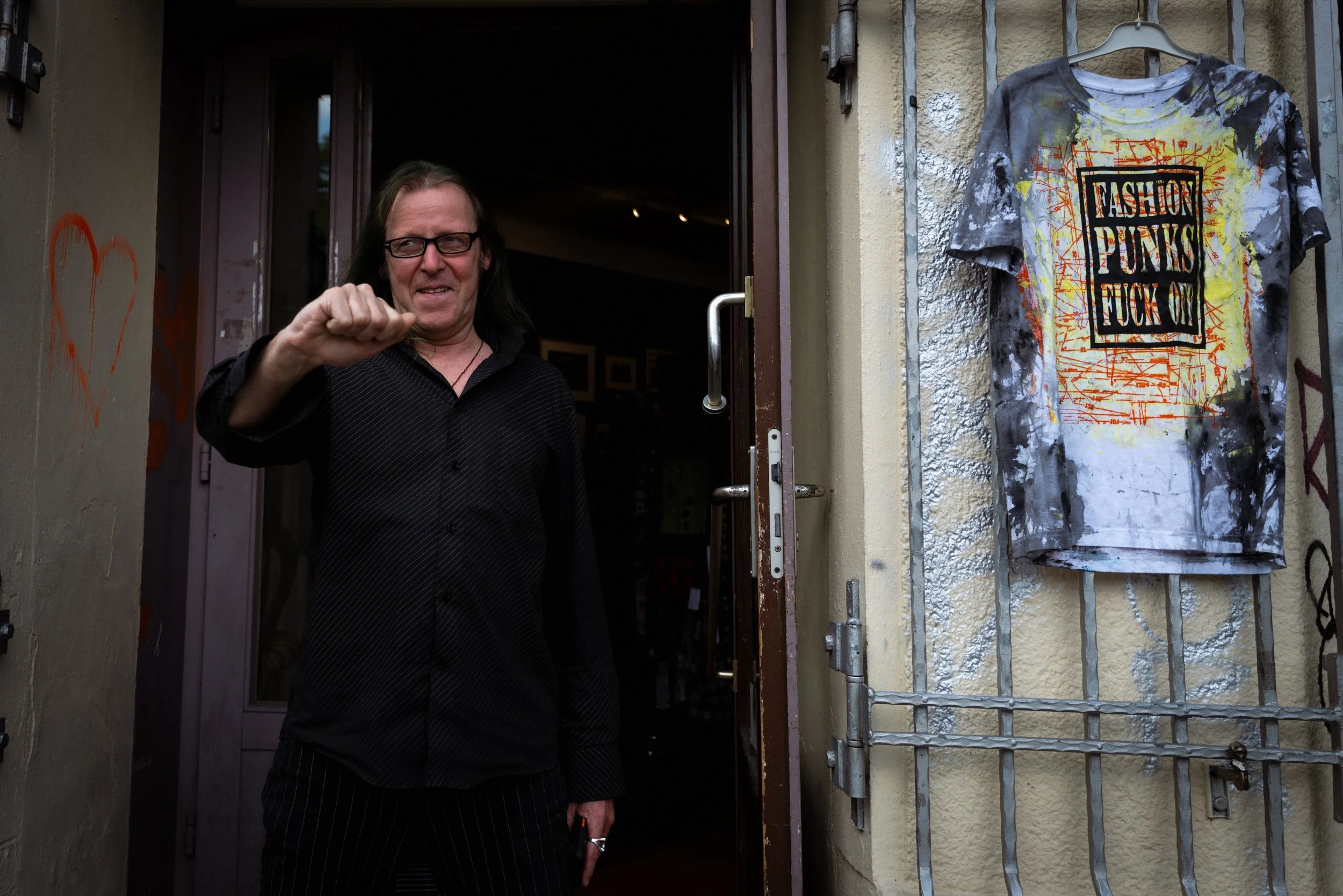 A man with long dark hair and glasses standing at an open door, with graffiti and a T-shirt hanging on a metal grid on the wall beside him. The T-shirt has paint splatters and the words 'Fashion Punks Fuck Off' printed on it.