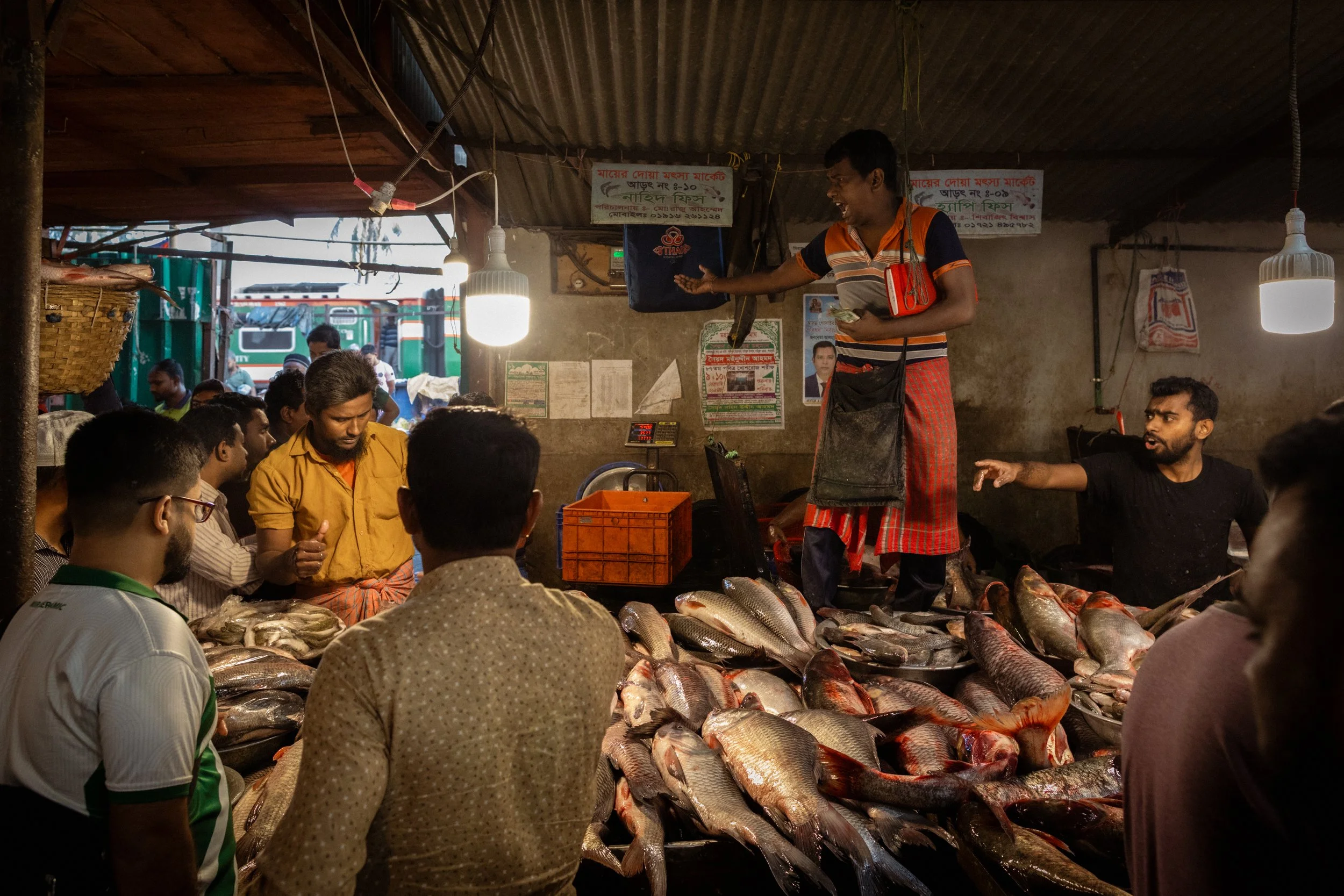 Fish vendor standing on a platform with fish displayed at a fish market stall, customers gathered around, some inspecting and purchasing fish, indoor setting with hanging lights and posters on the wall.