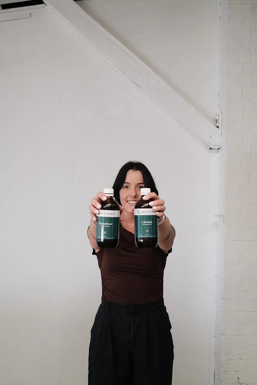 A woman with black hair smiling and holding two bottles of supplements or vitamins, one labeled Passionflower and the other Calendula, in front of a plain white wall.