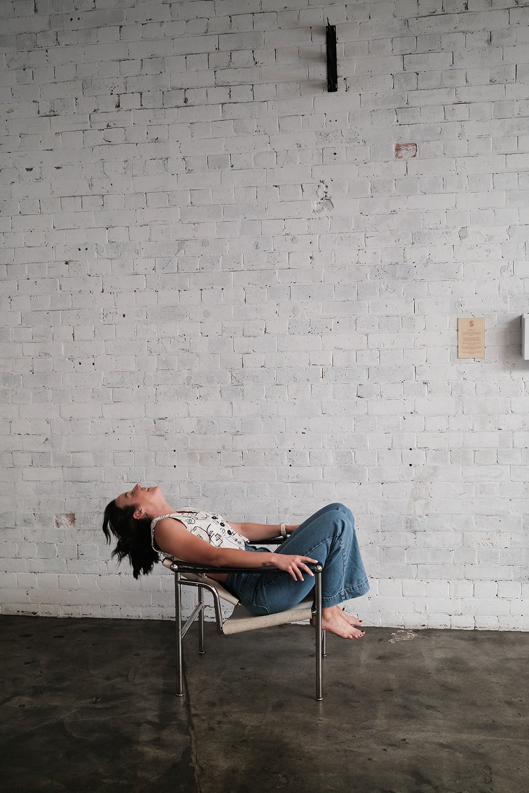 A woman with dark hair, wearing a white patterned sleeveless top and blue jeans, sits on a modern chair with her head tilted back and eyes closed, barefoot in a room with a white brick wall and a concrete floor.