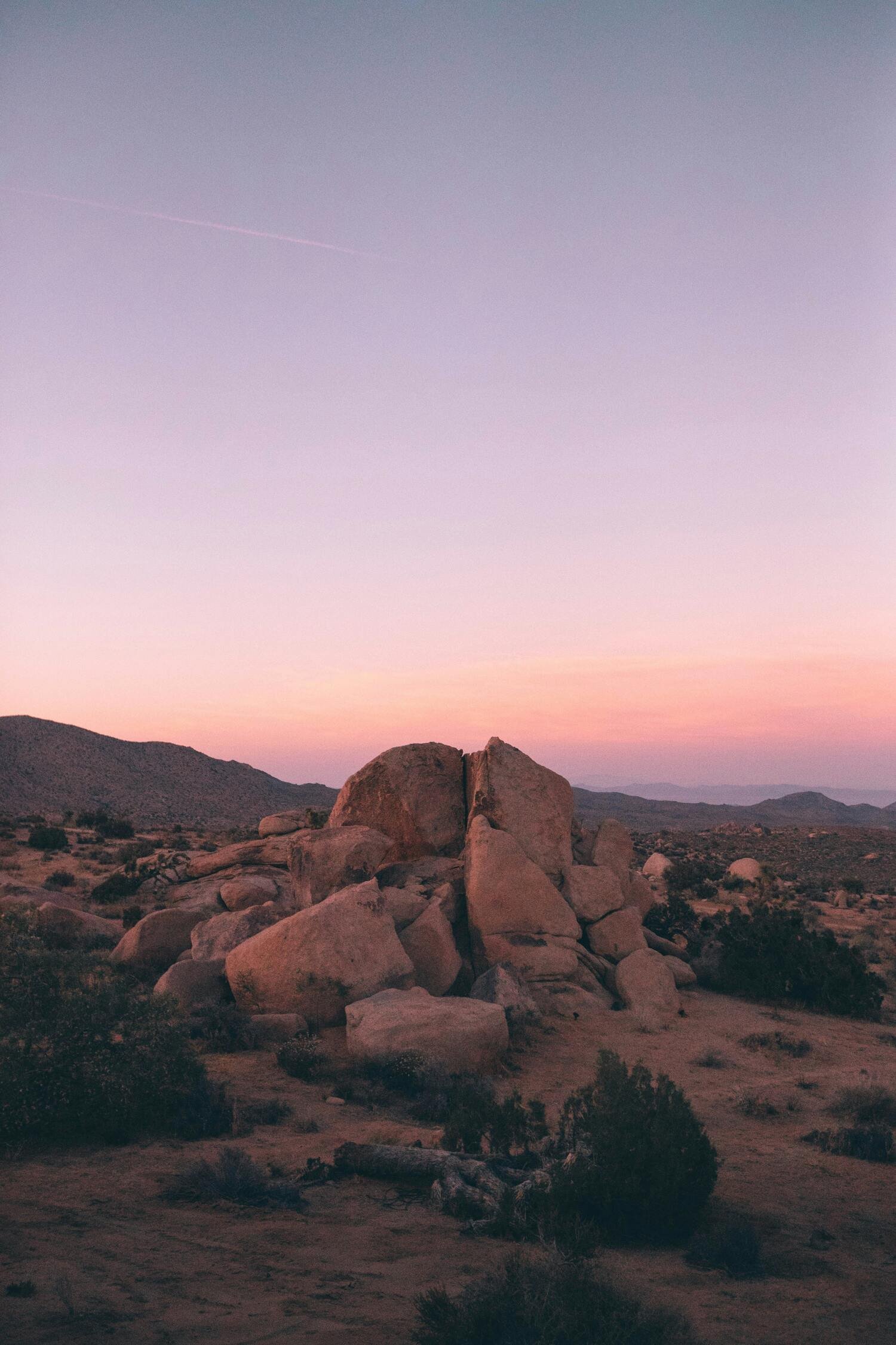 A landscape view of a desert with large boulders, sparse bushes, and distant mountains under a pastel pink and purple sky at sunset.