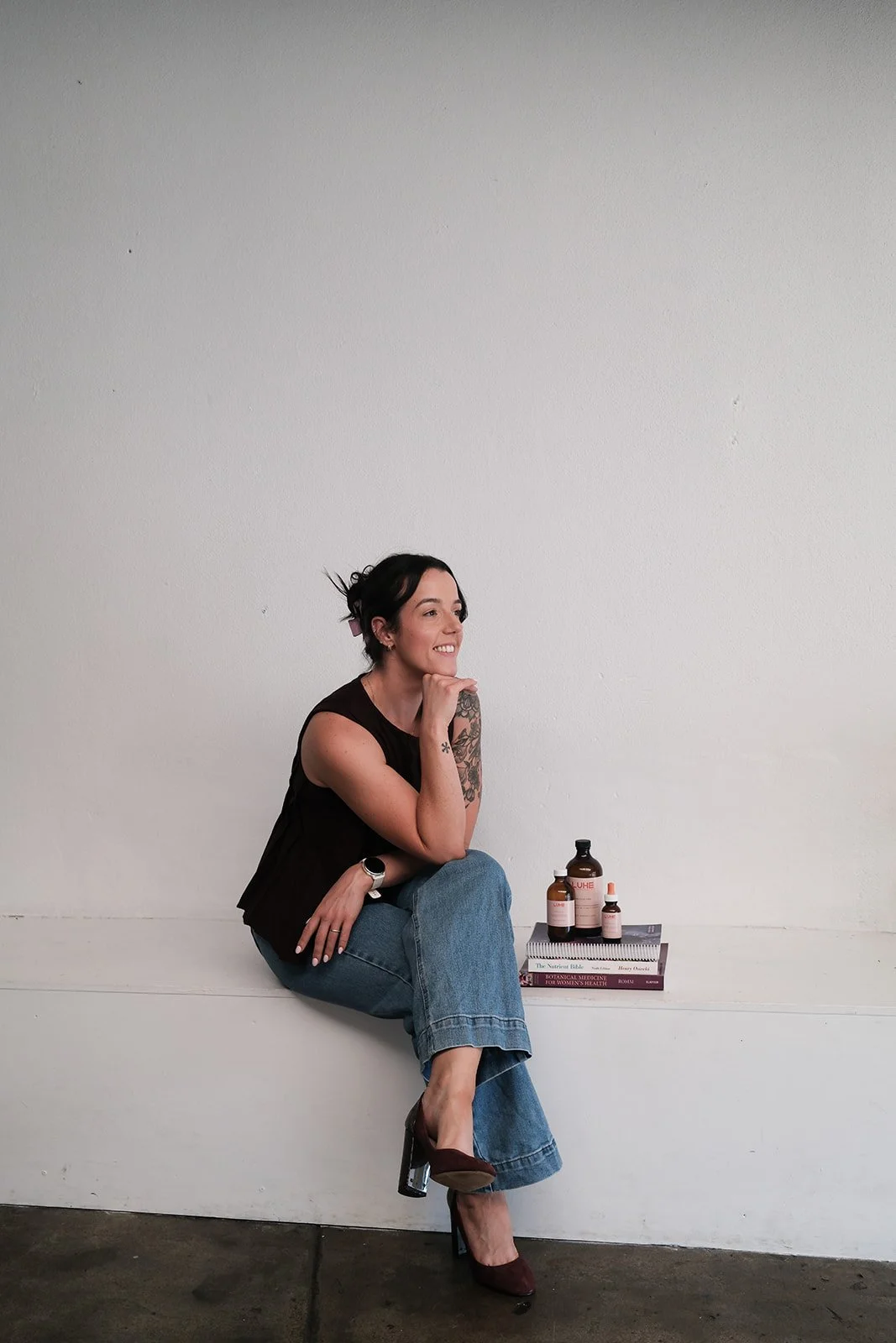 Clancy, a naturopathy, a woman with dark hair sitting on a white bench against a plain white wall, smiling, with a stack of books and several bottles on top of the books beside her.