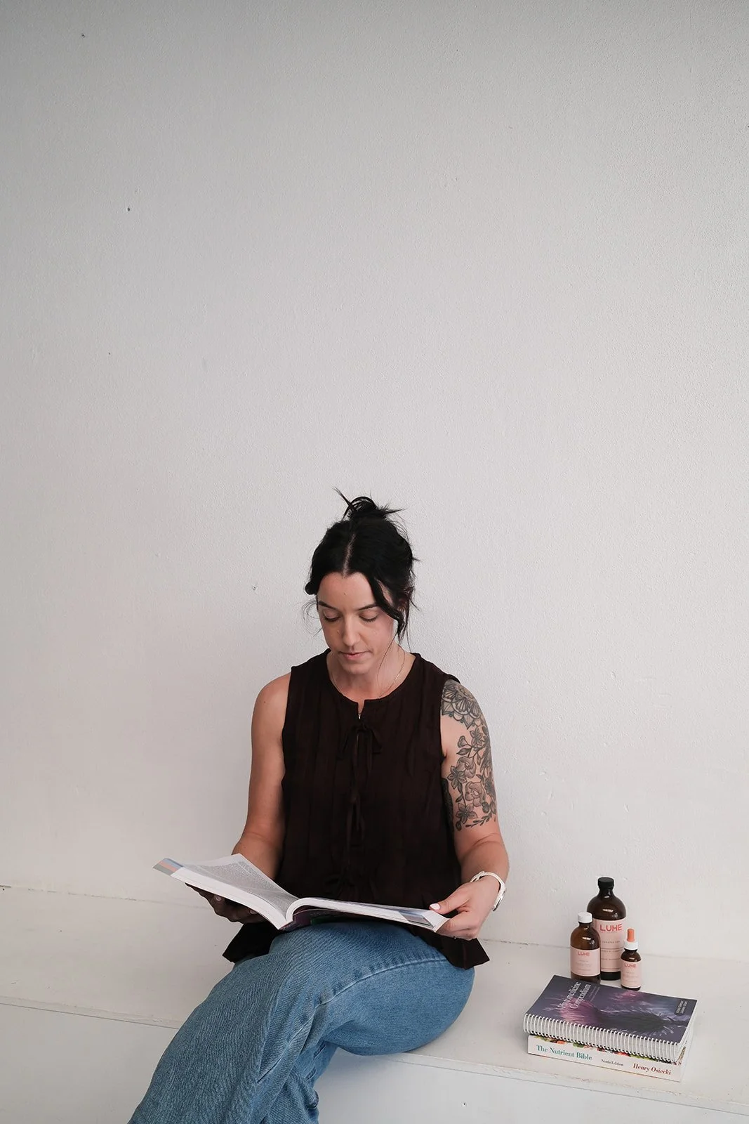 A woman with a tattoo on her right arm, wearing a black sleeveless top and blue jeans, sitting cross-legged against a plain white wall, reading a book. Beside her on the floor are several bottles and a spiral-bound book.