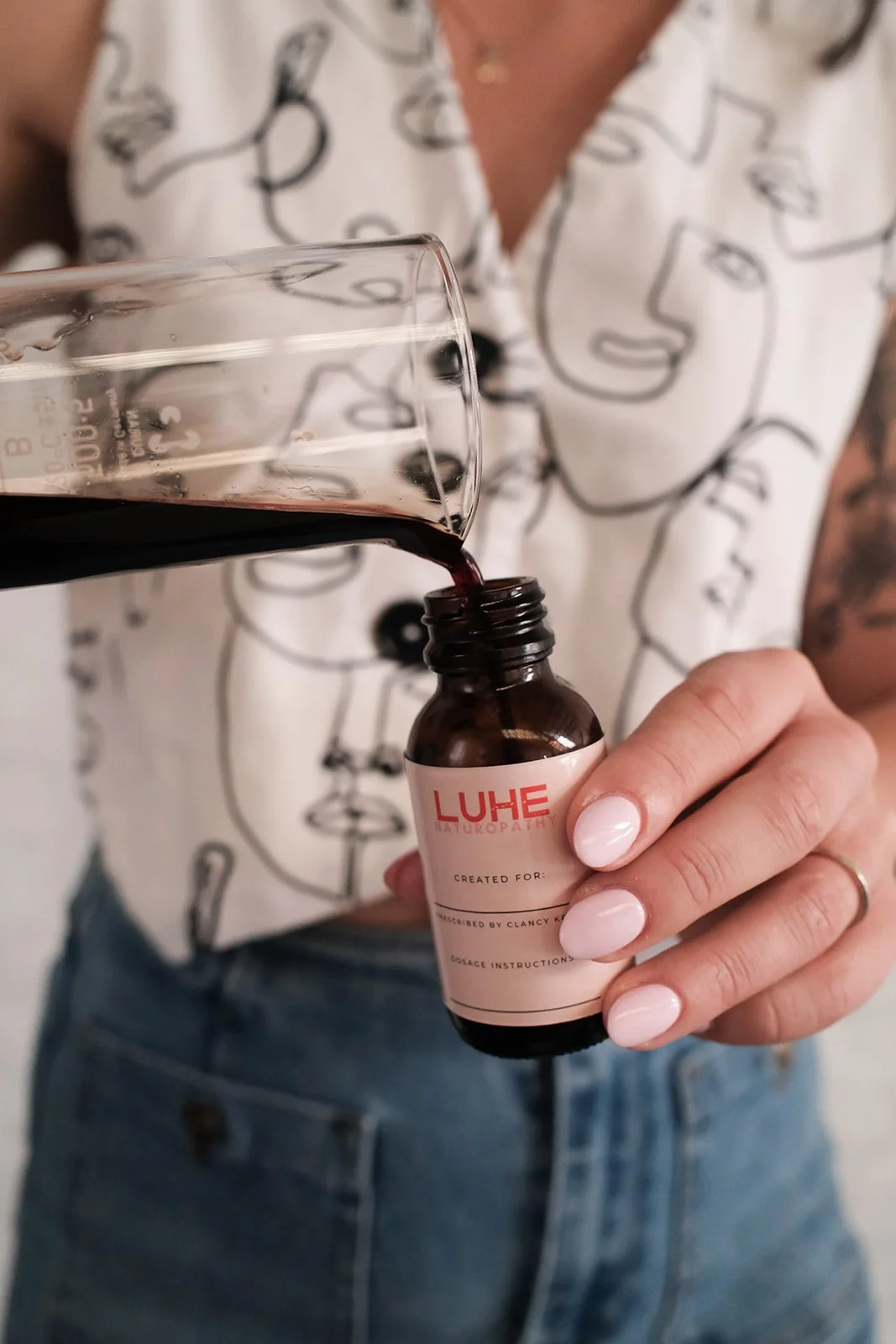 A person with pink nail polish holding a small brown bottle labeled "LUHE" and pouring liquid herbs into a glass bottle.