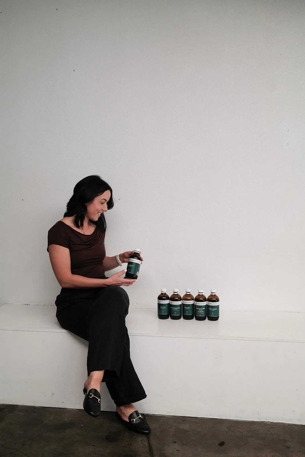 A female naturopathy, Clancy, sitting on a white bench against a plain wall, holding a bottle of herbs and surrounded by additional bottles on the ground.
