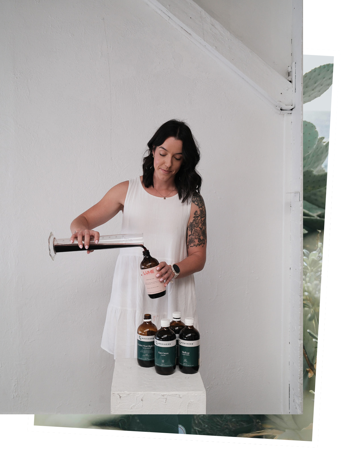 A naturopathy, Clancy, a woman in white dress pouring liquid herbs into a bottle from a graduated cylinder, with three other bottles on a white block.