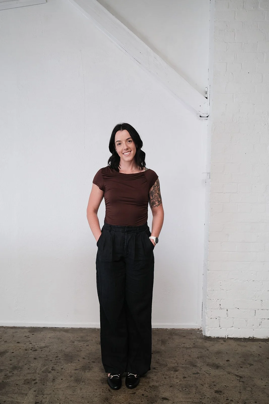 A woman with shoulder-length black hair, wearing a brown top, black pants, and black shoes, standing against a white wall with a brick corner.