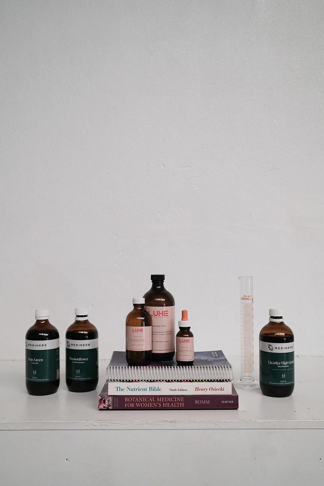 Arrangement of herbal medicine bottles, a graduated cylinder, and books on a white surface against a plain white wall.