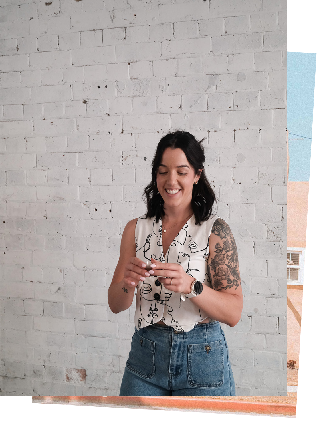 A woman with dark hair and tattoos on her right arm, smiling and looking at a shot of liquid herbs, standing against a white brick wall.