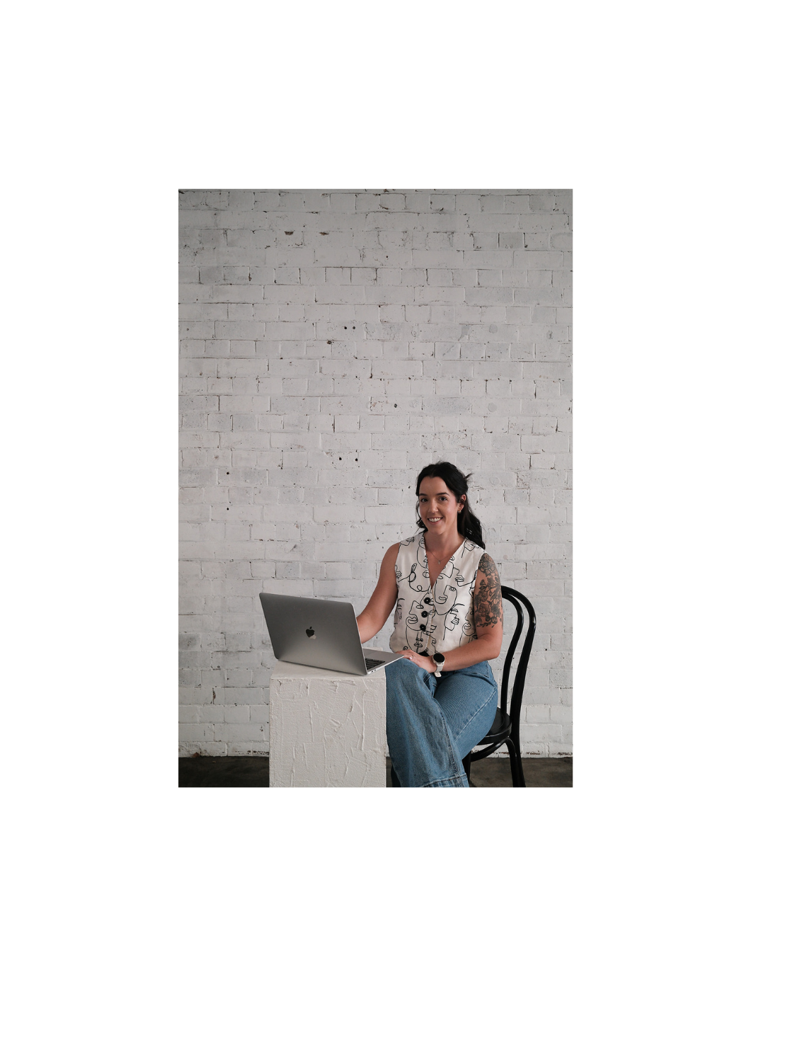 A naturopathy sitting at a white pedestal with a silver MacBook laptop, smiling, against a white brick wall background.