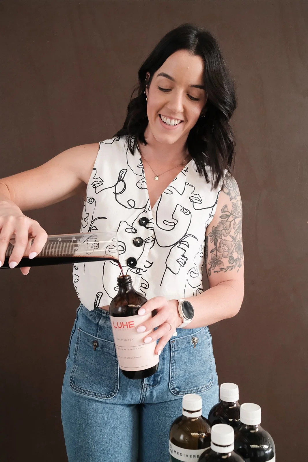 Woman with dark hair, wearing a sleeveless shirt with abstract face designs, pouring liquid herbs into a bottle from a graduated container. She has tattoos on her right arm, a watch, and is smiling. Several bottles are on the table in front of her.
