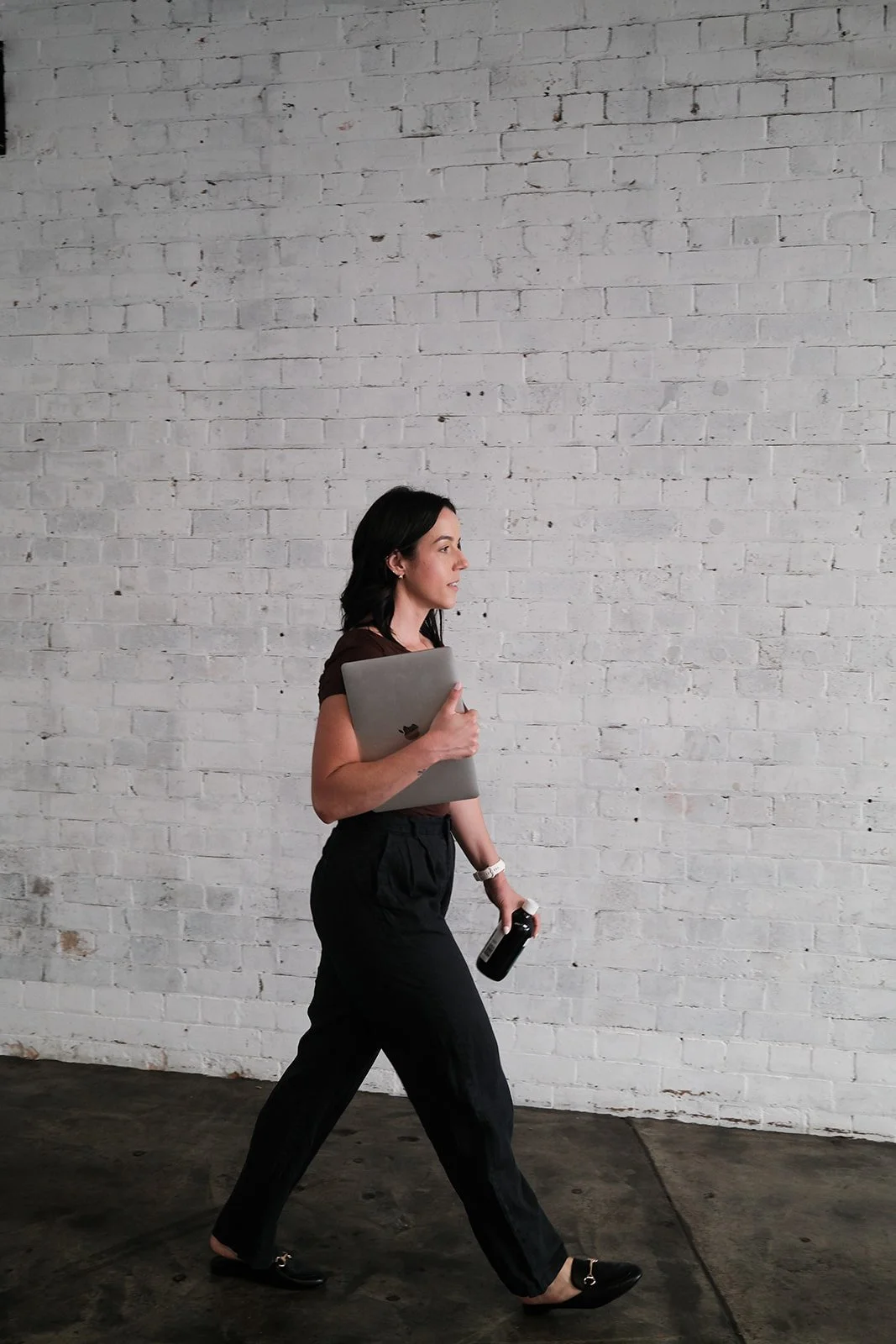 A woman walks past a white brick wall, holding a laptop and a bottle in her hands.