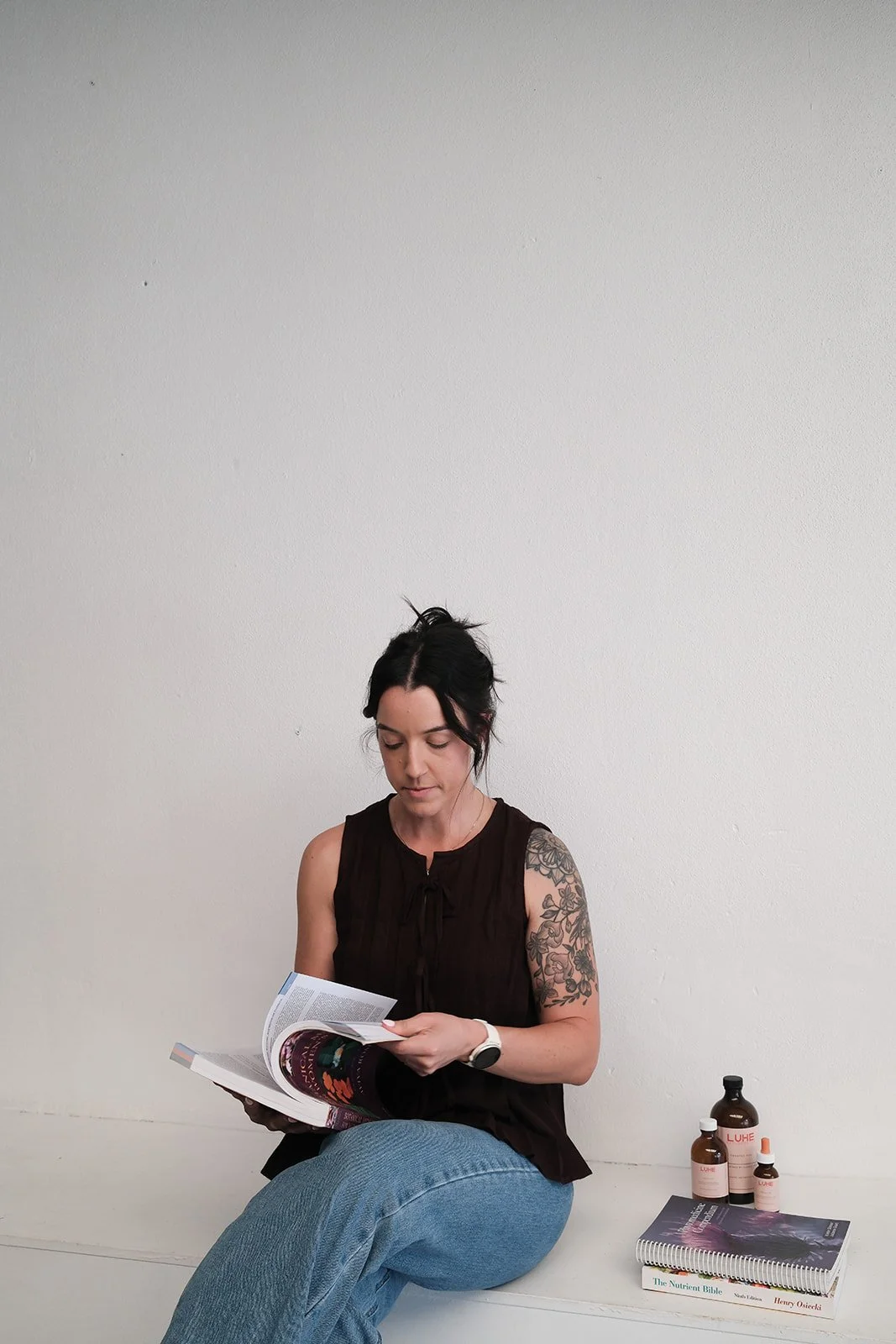 A woman with dark hair, dressed in a sleeveless black top and light blue jeans, sitting against a plain white wall, reading a magazine. Beside her are bottles and dropper bottles labeled 'LUHE' and a few books, including 'The Nutrient Bible' and others.
