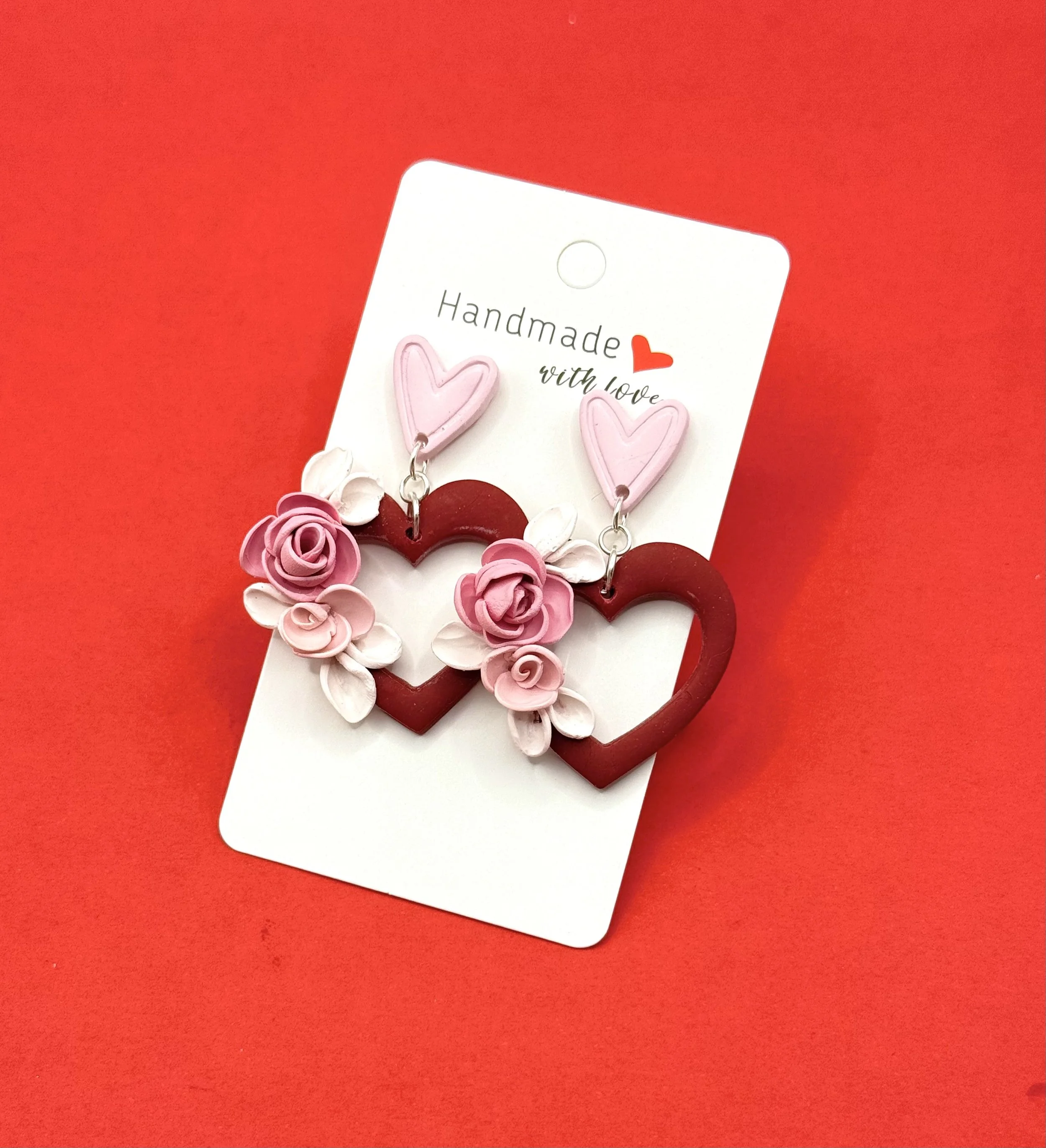 Pair of handmade heart-shaped earrings with pink and red roses, displayed on a white card against a red background.