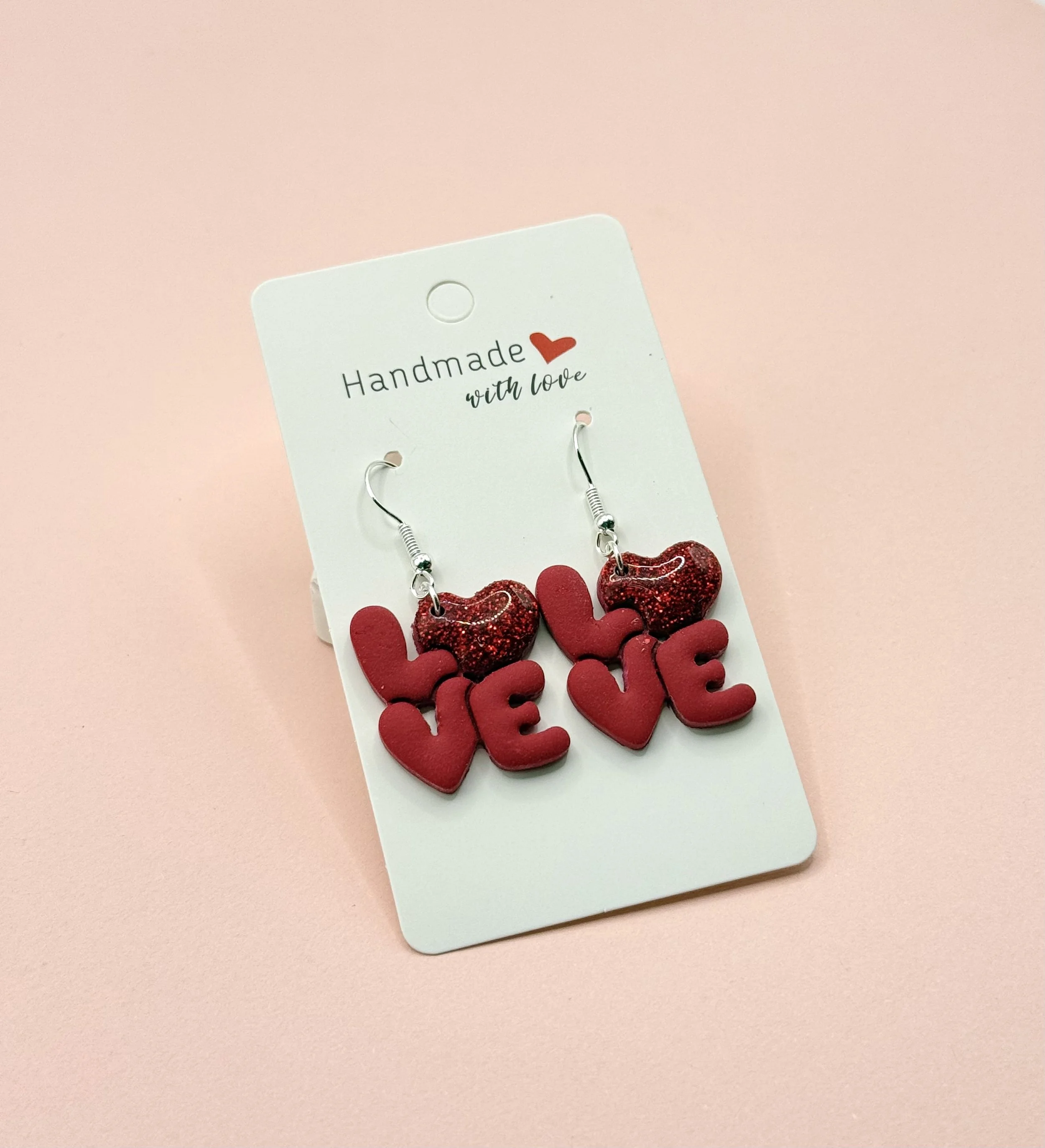 Pair of handmade earrings with red glitter hearts and red letters spelling 'LOVE' on a white card against a pink background