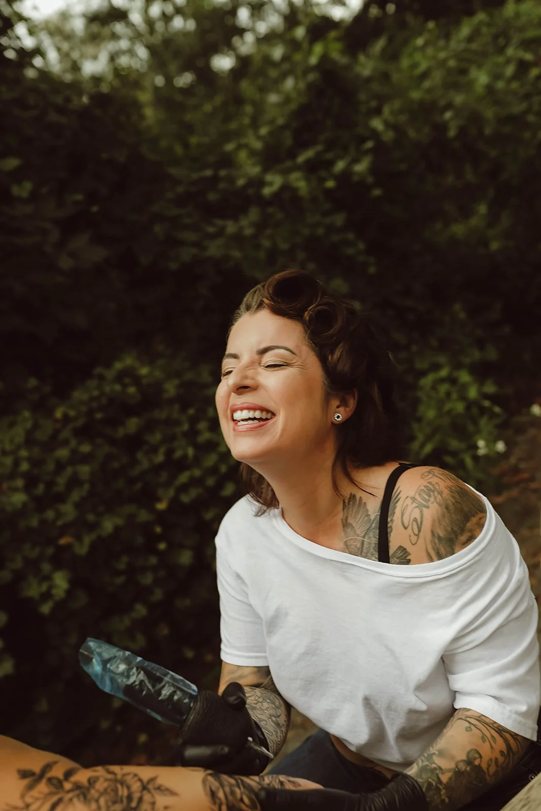 A woman with tattoos smiling outdoors against a leafy background, holding a tattoo machine.