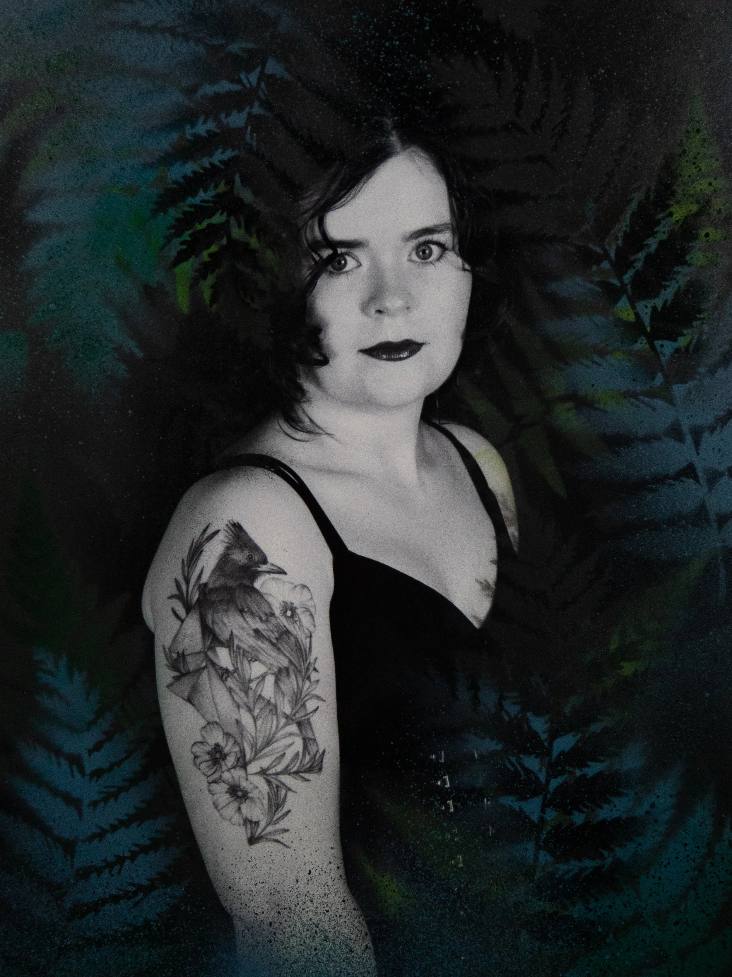 Black and white portrait of a woman with dark hair and dark lipstick, surrounded by green and blue fern leaves in the background. She has a floral and bird tattoo on her upper arm.