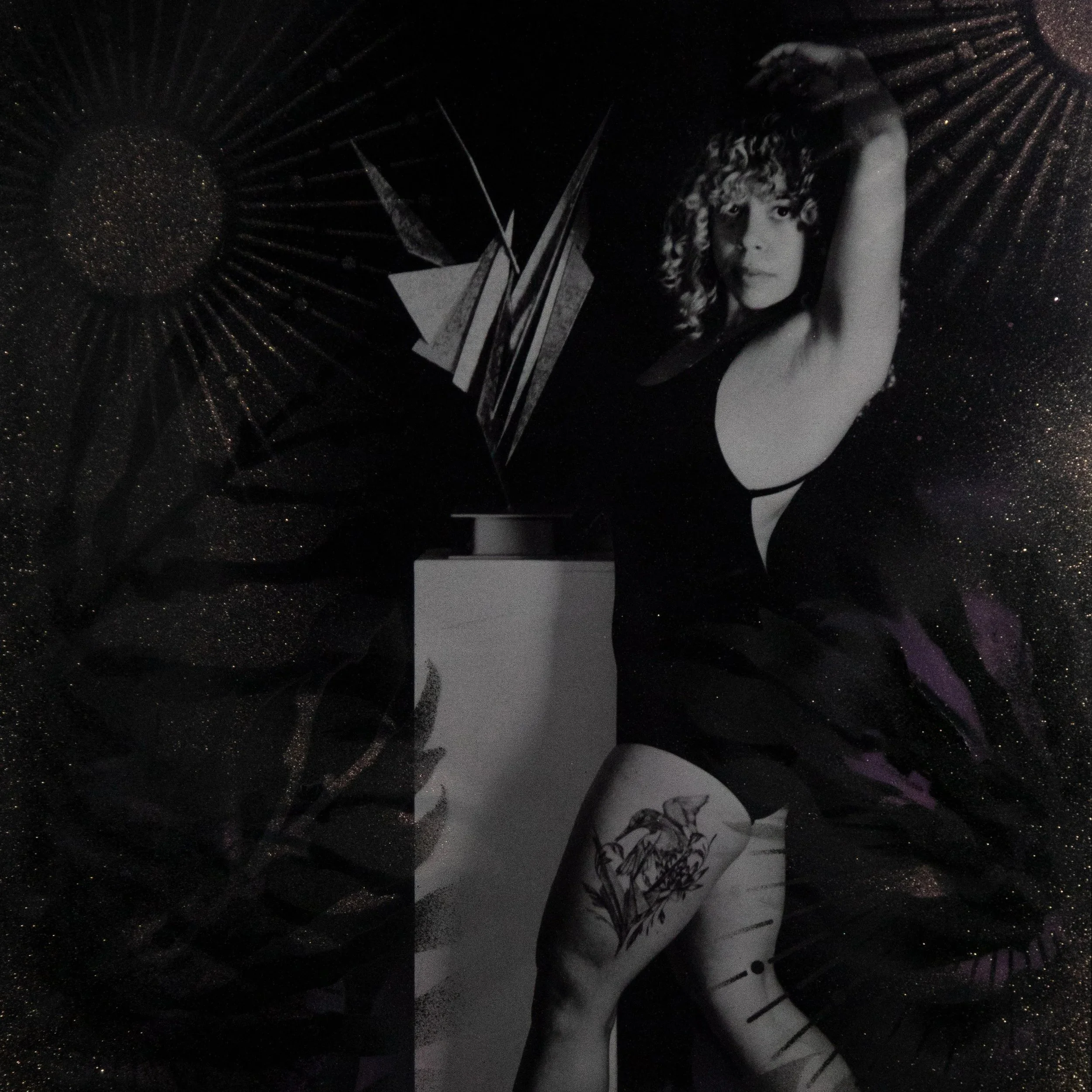 A woman with curly hair in a black dress and fishnet stockings with a tattoo on her thigh, standing next to an abstract sculpture in a dark, glittery setting.
