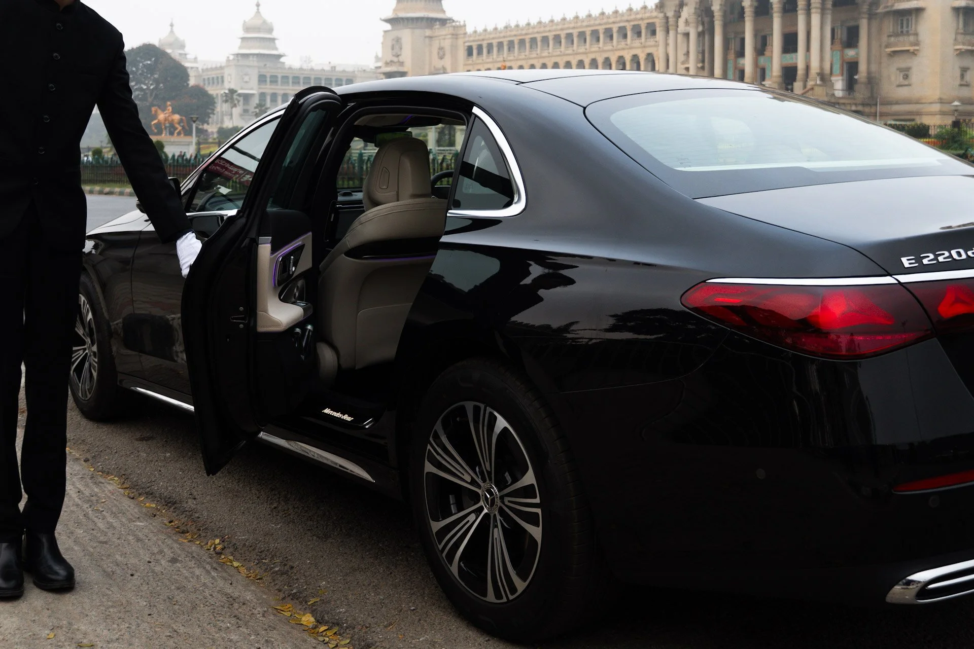 A black luxury sedan with its rear door being opened by a professional chauffeur with white gloves, parked on a city street in Bangalore.