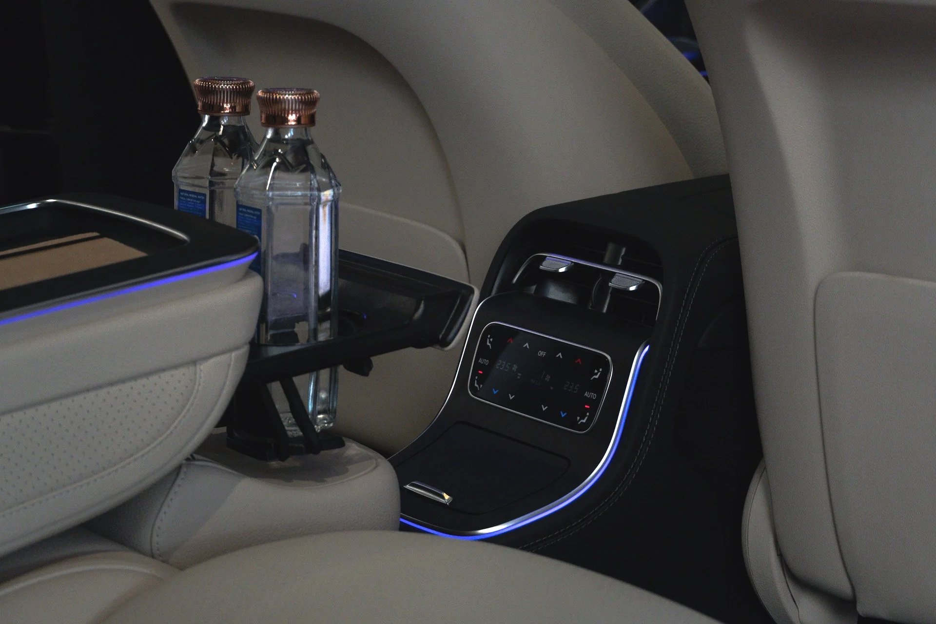 Car interior with a rear seat console holding three water bottles, climate control panel with digital display and ambient lighting.