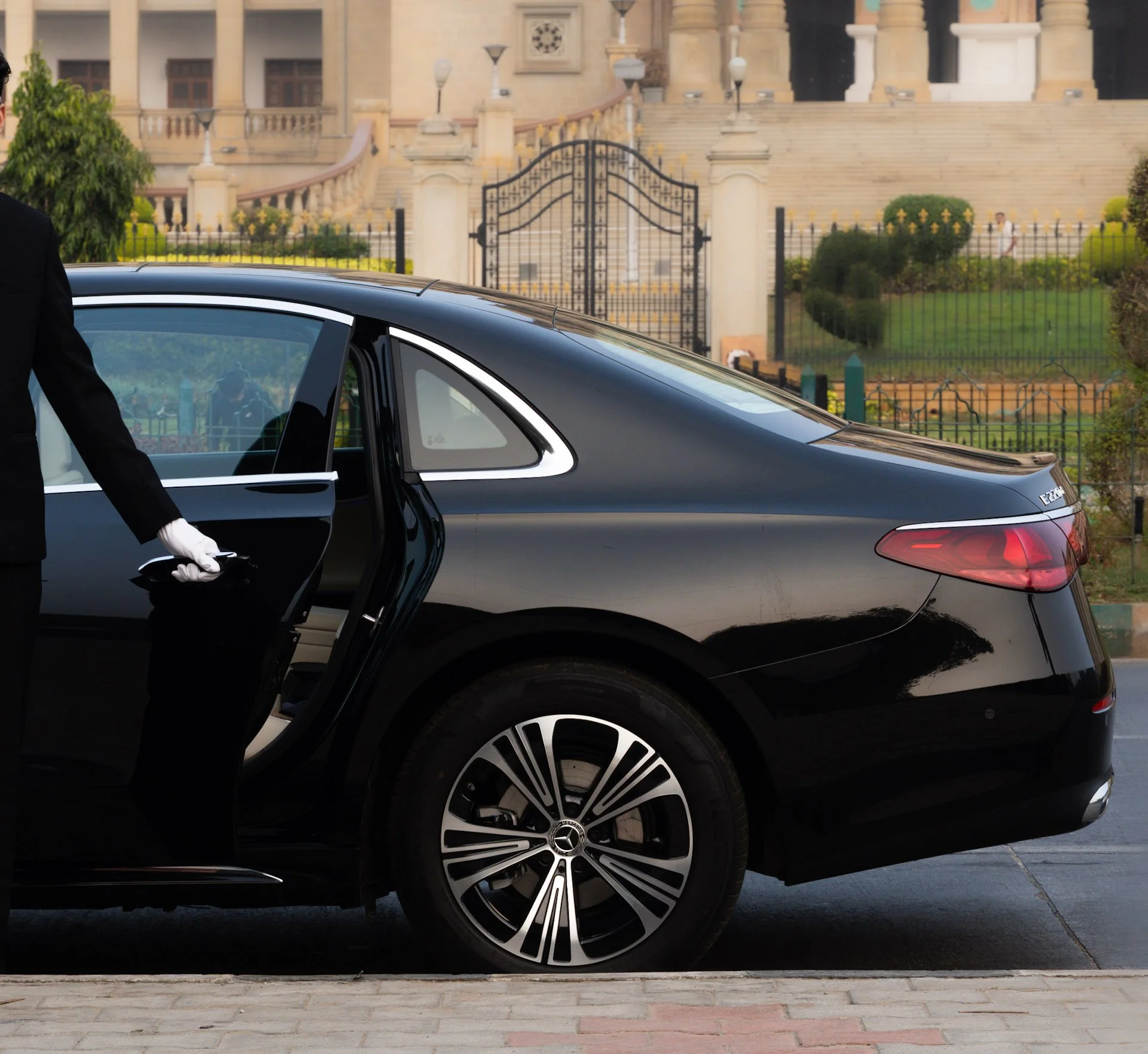 A black luxury car with its rear passenger door open, a chauffeur in a black suit and white gloves is opening the door. The vehicle is parked on a paved area near a building with grand steps and a wrought iron gate, surrounded by greenery.
