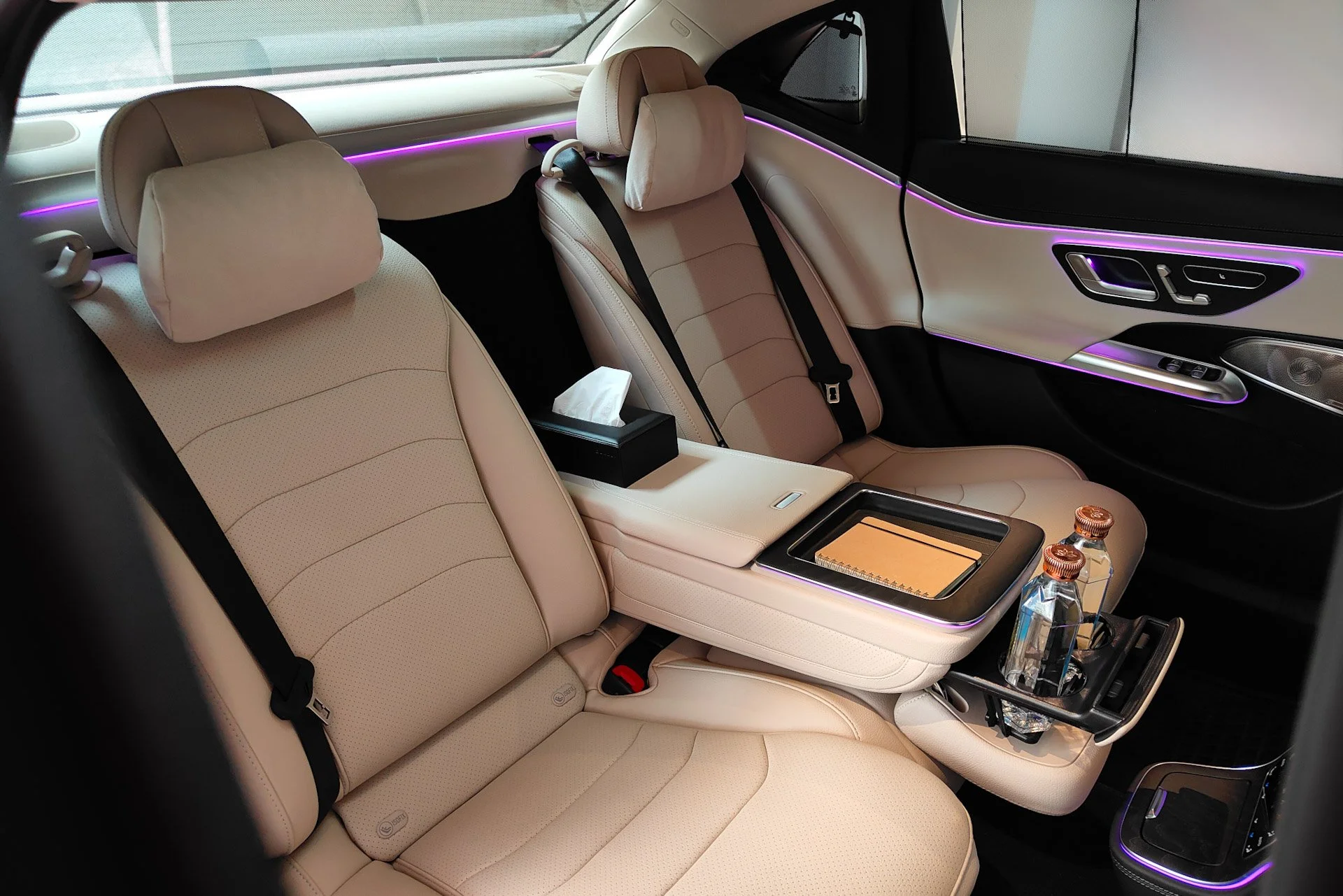 Luxury car interior with beige leather seats, purple ambient lighting, a tissue box, a notebook, two water bottles, and control buttons on the door panels.