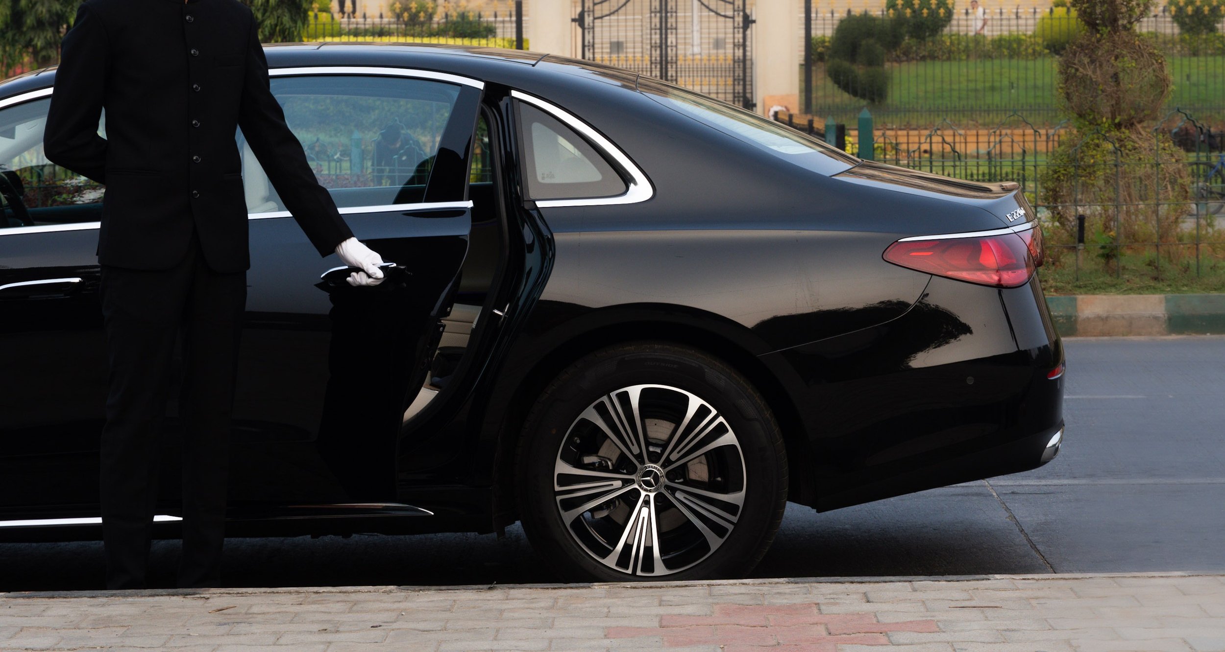 A chauffeur in formal attire with white gloves is standing next to a black luxury sedan with the rear passenger door open.