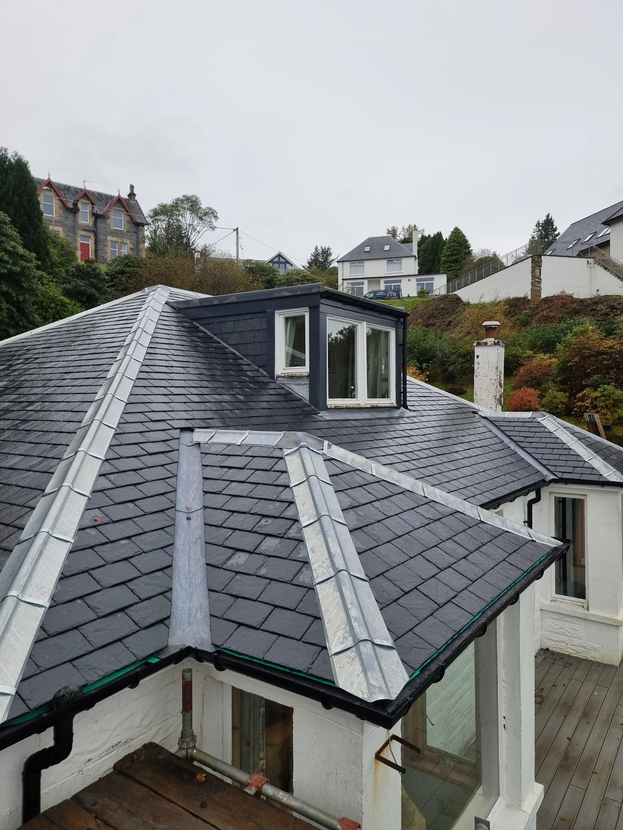 View of a house roof with dark gray tiles and a dormer window, overlooking neighboring houses and a hillside with trees and plants, in an overcast sky.