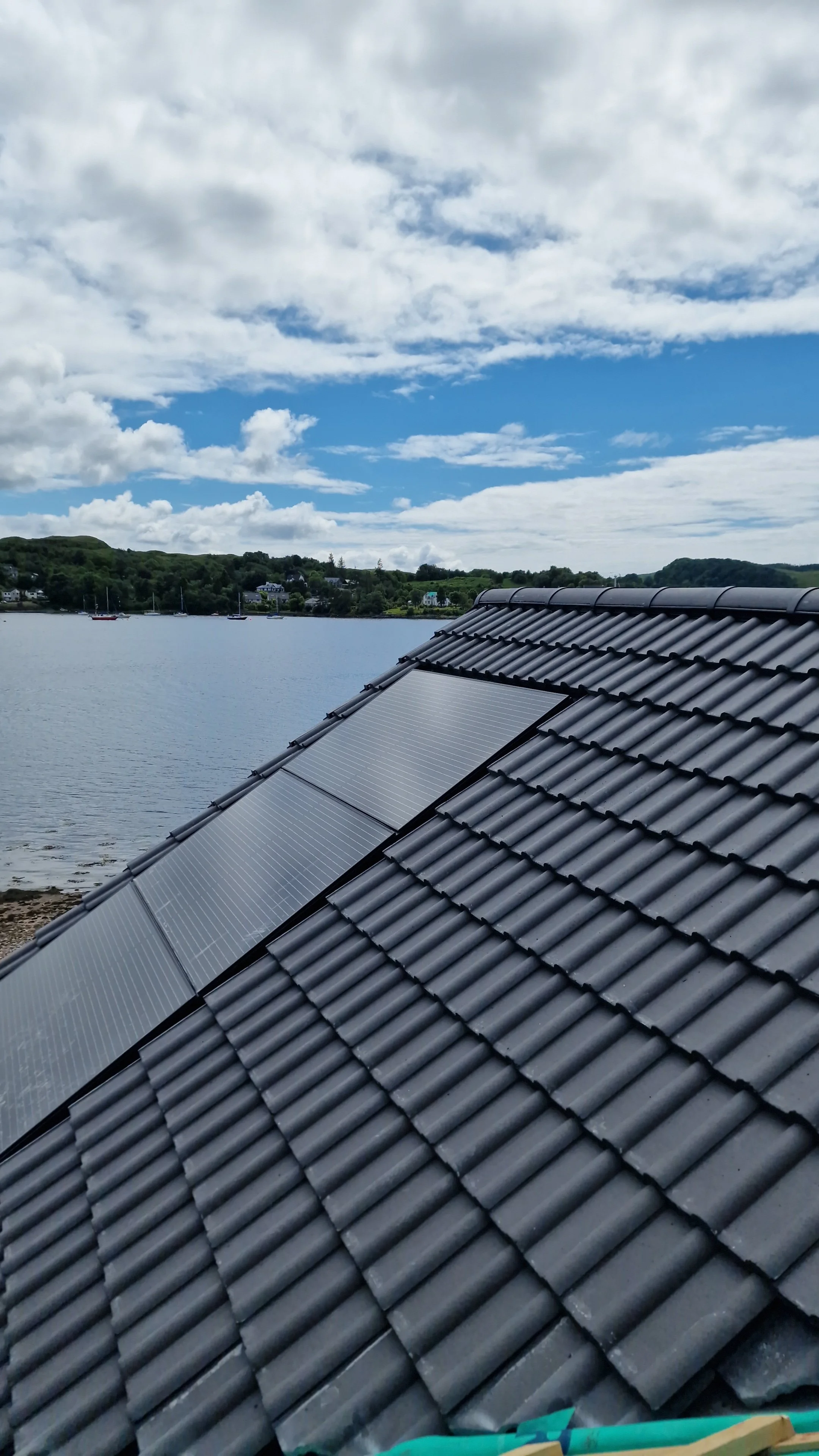 Black tiled roof with solar panels overlooking a body of water and cloudy sky.