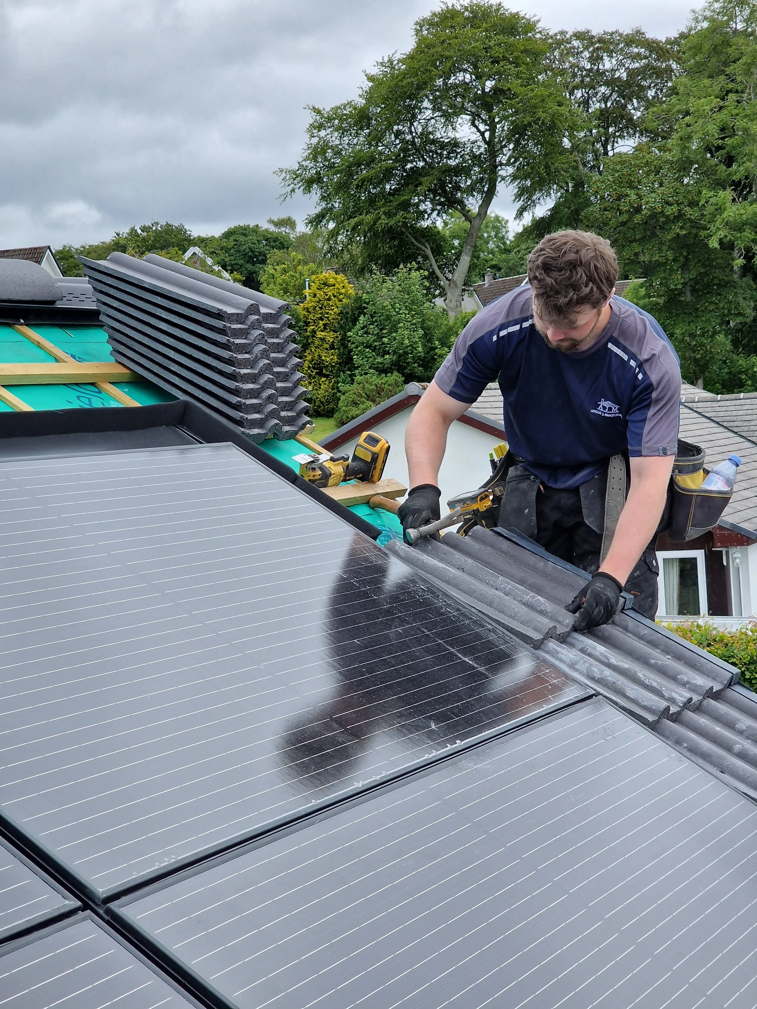 A man installing solar panels on a roof. He is wearing gloves and using a power tool, with a toolbox and other tools nearby. The roof is partly covered with new solar panels, with some roofing material visible in the background. Trees and houses are 