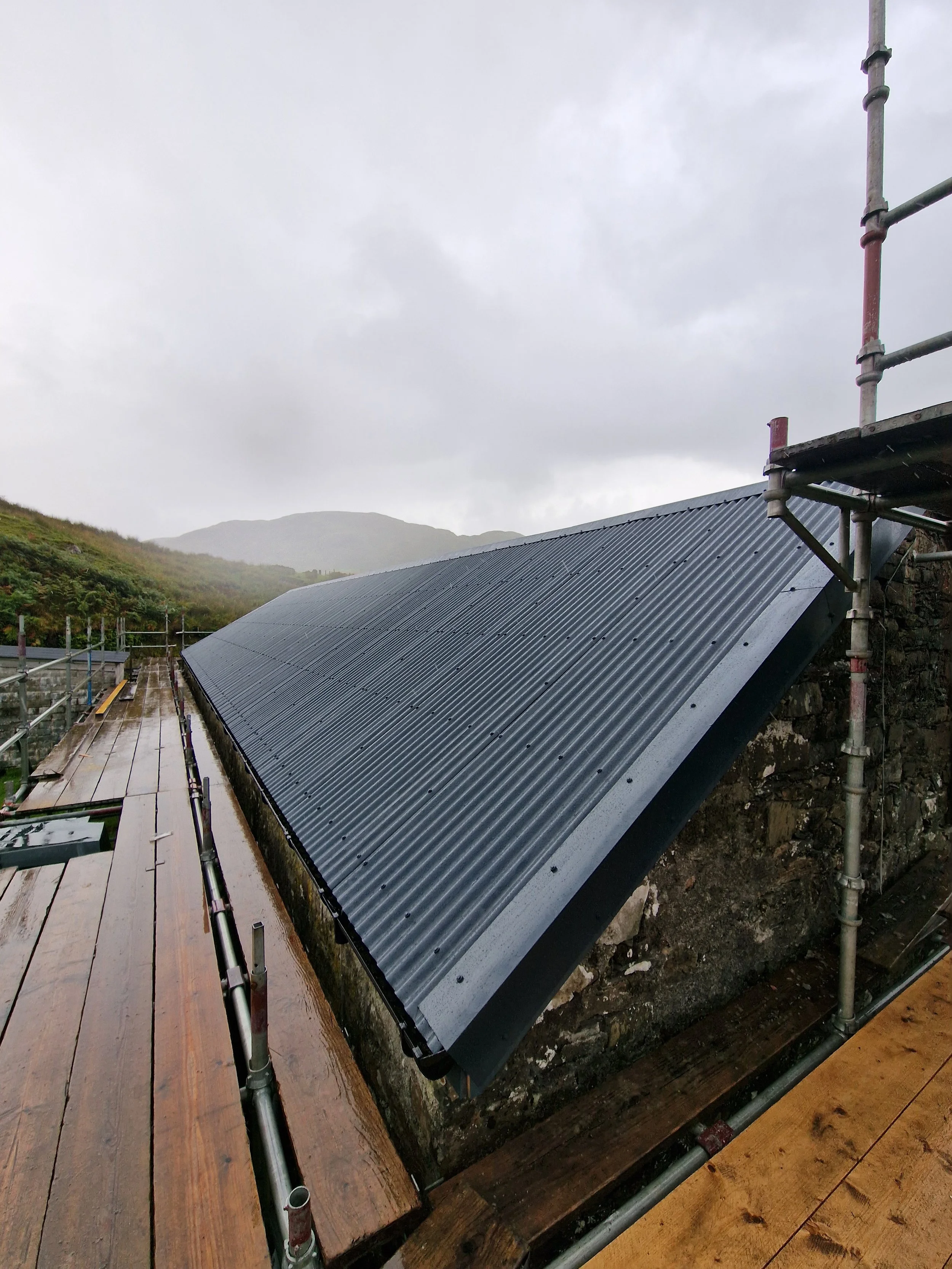 A metal roof installation on a building situated on a hillside, with scaffolding and wet wooden planks nearby, under cloudy and rainy weather.