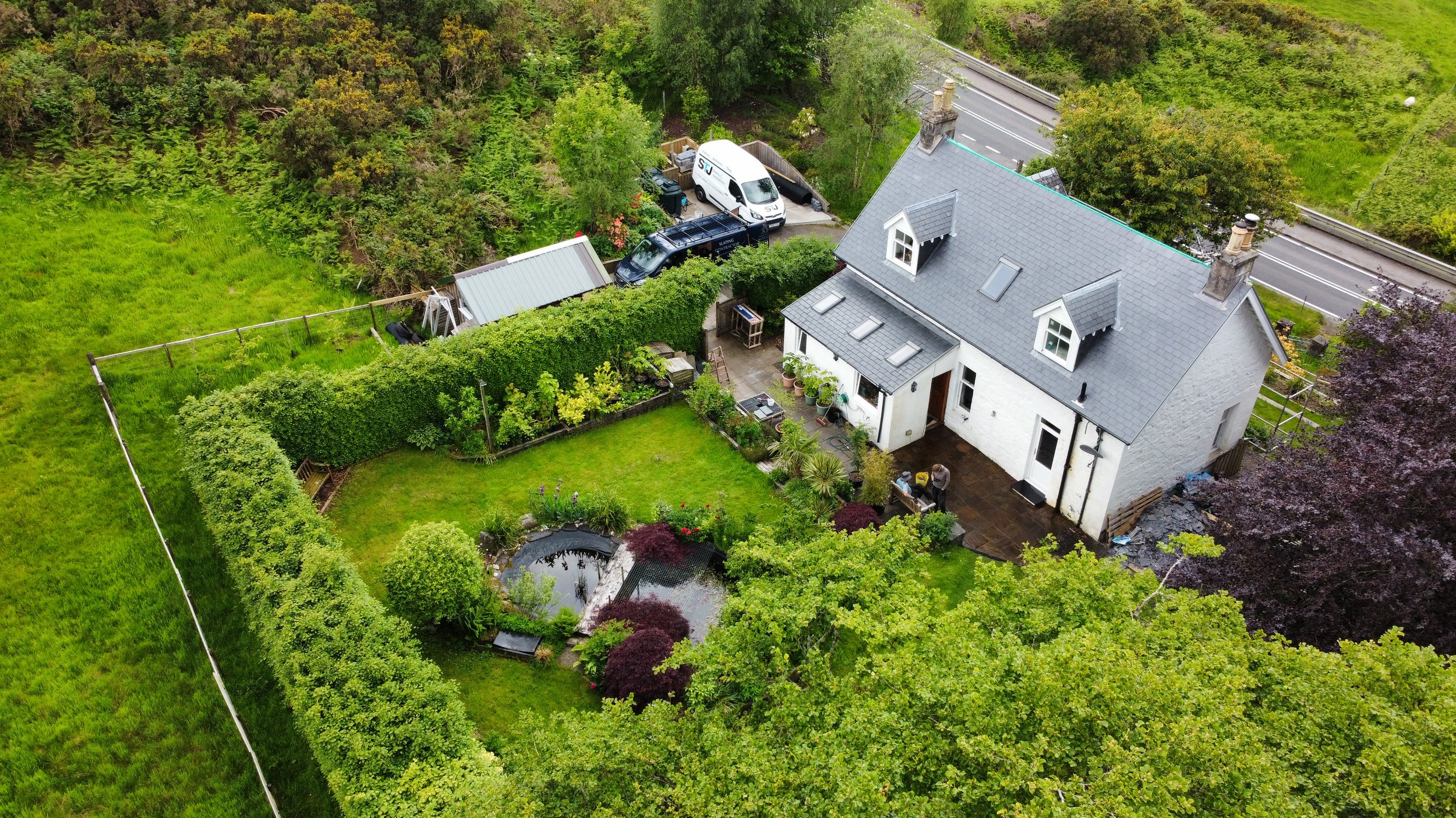 Aerial view of a white house with a gray roof, surrounded by a lush green garden with trees and bushes, a small pond, and a patio area with potted plants. A driveway with parked vehicles is visible behind the house, and a road runs along the top of t