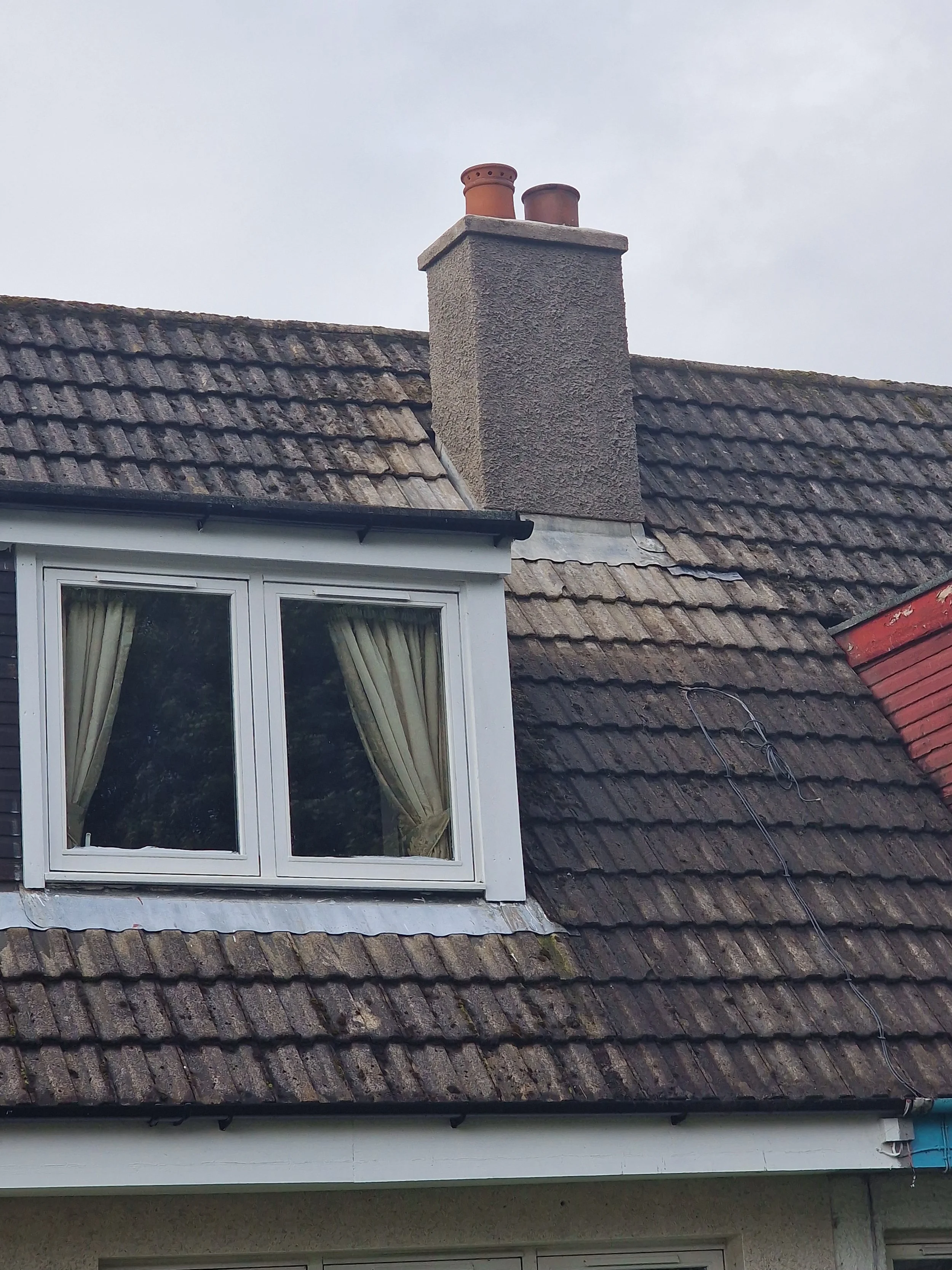 Close-up of a house roof showing a large chimney and a window with yellow curtains. The roof has aged, mossy tiles, with a section of red tiles and some black wires visible.