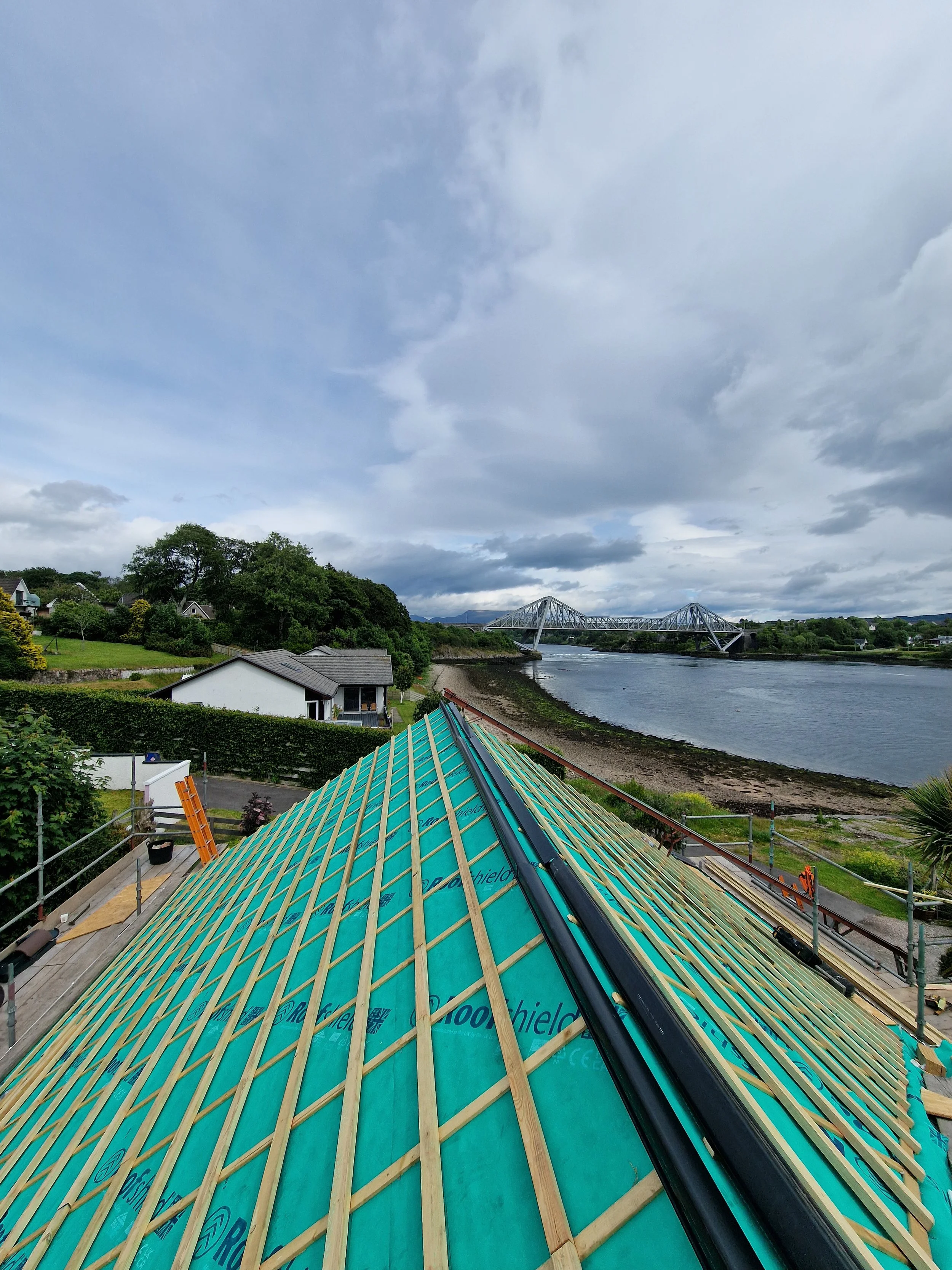 Under construction roof with wooden beams and a black gutter, overlooking a river with a bridge and cloudy sky.