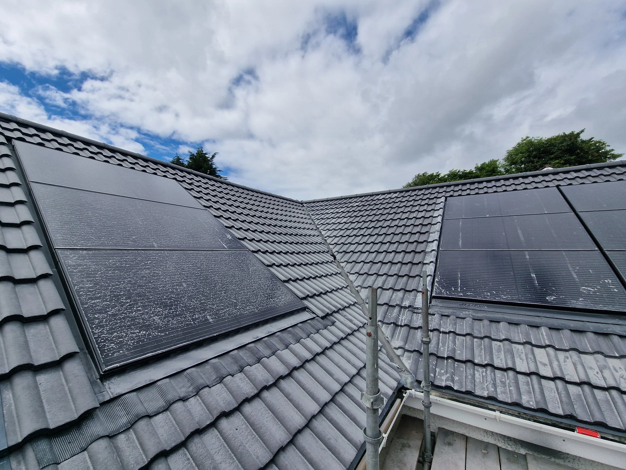 Residential roof with new solar panels installed on gray tiles, under partly cloudy sky, with trees in background.