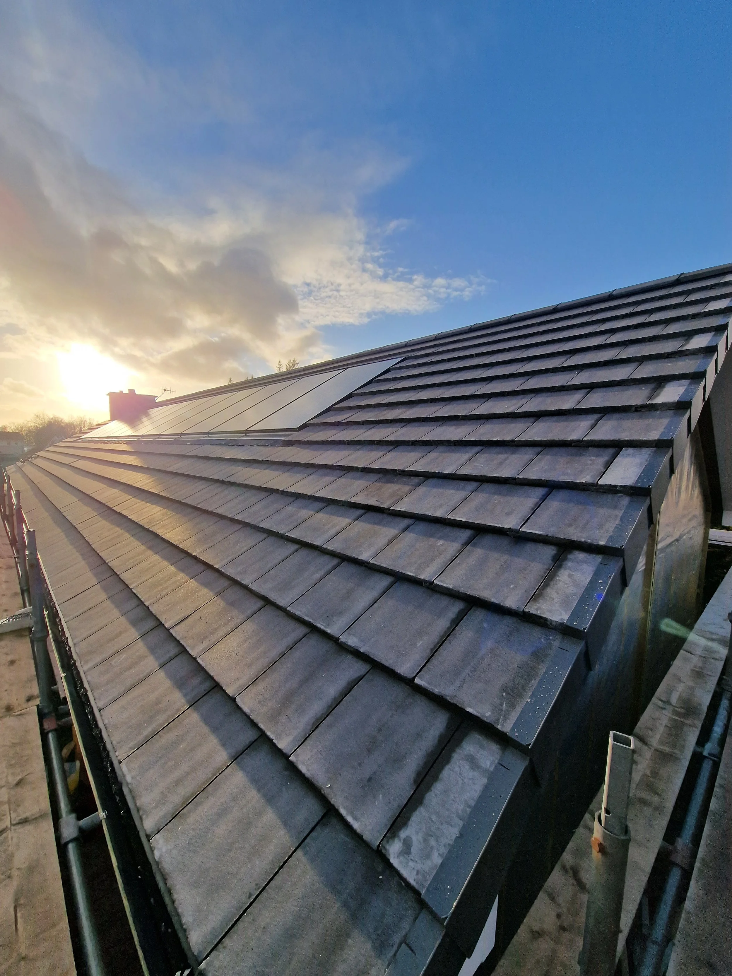 View of a house roof with black tiles and solar panels installed, taken during sunset.