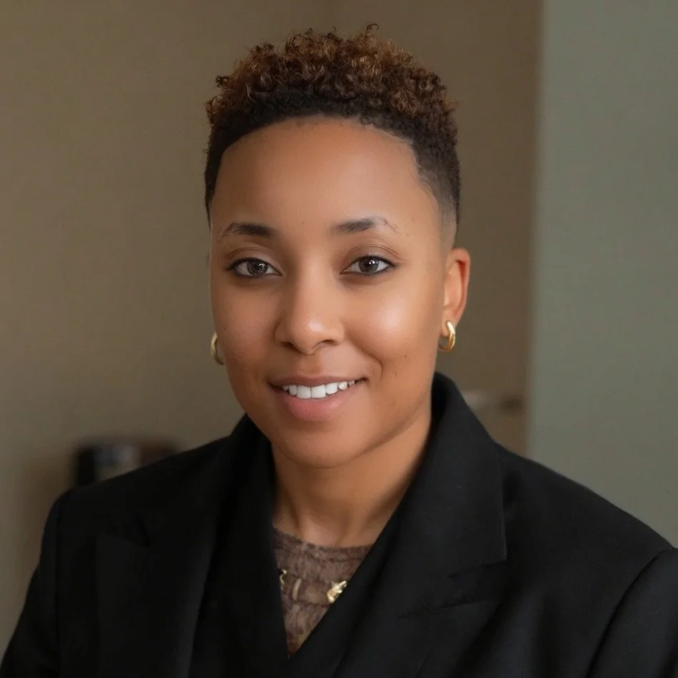 An African American woman with short curly hair, gold hoop earrings, and a black blazer smiling at the camera in an indoor setting.