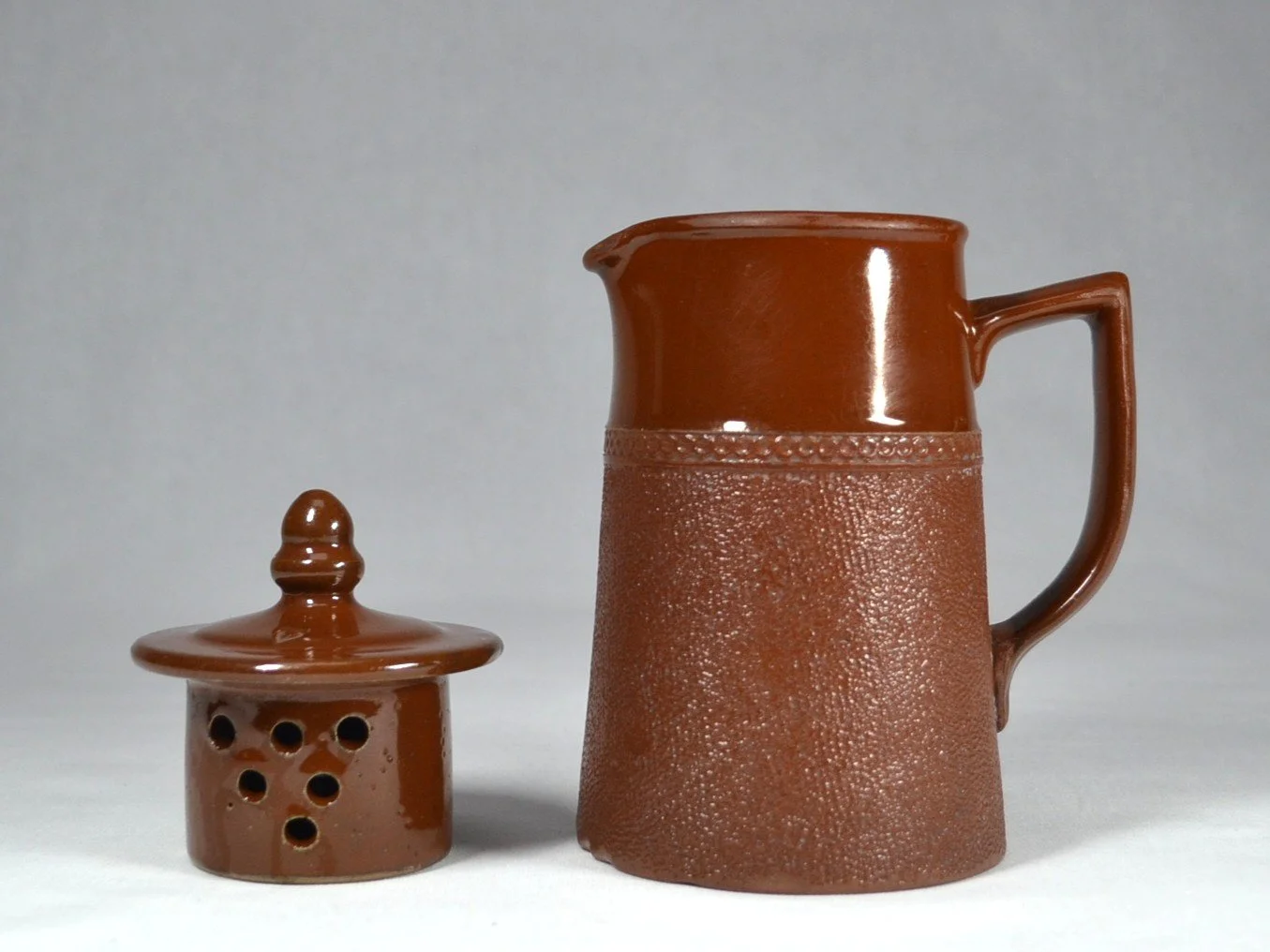 A brown ceramic pitcher with a textured lower half and a smooth upper half, accompanied by a small brown ceramic lidded container with holes and a rounded knob on top.