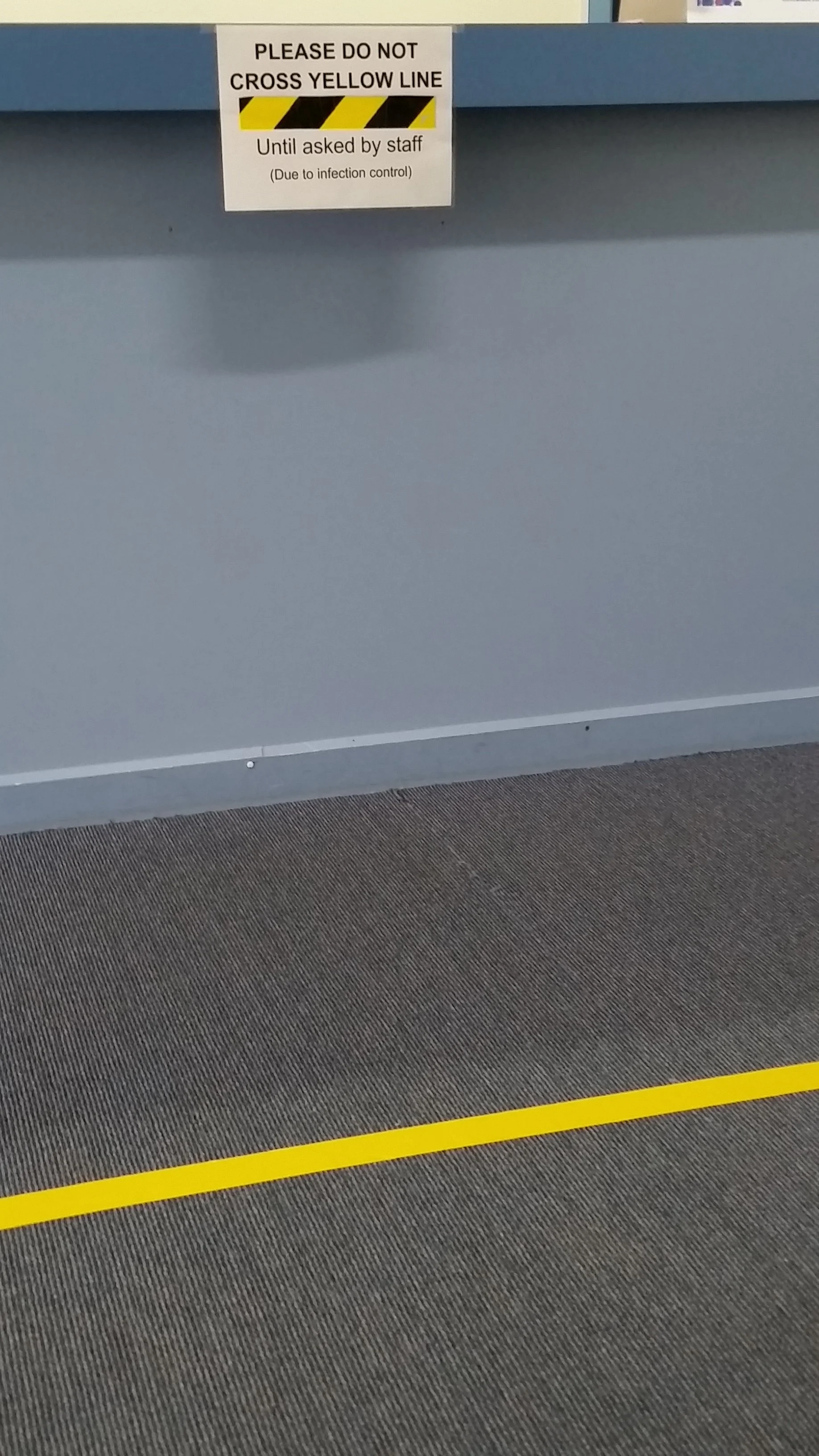 A sign on a blue surface that says, "Please do not cross yellow line until asked by staff (due to infection control)." There is a yellow and black striped line on the gray floor below the sign.