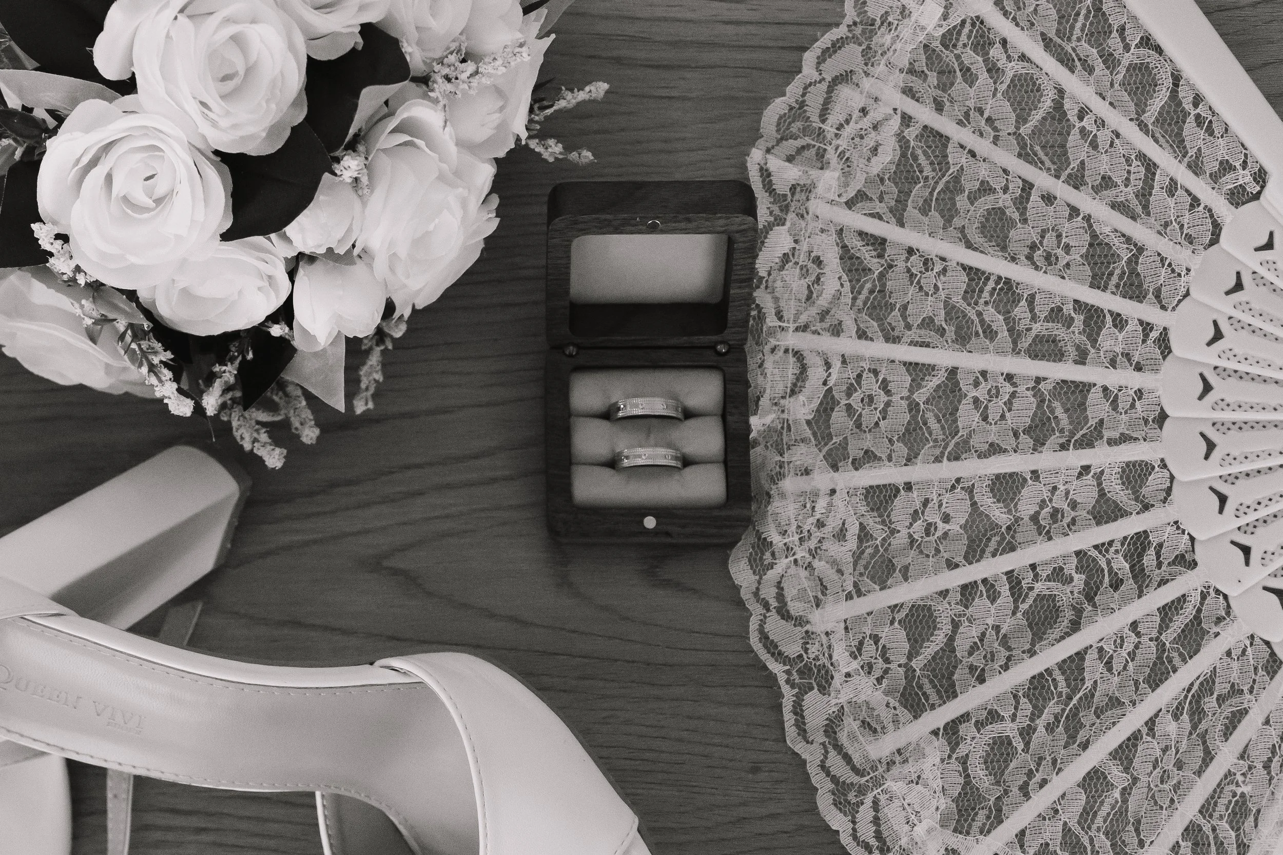 Ein offener Ringkarton mit zwei Eheringen, eine Blumenbouquet mit weißen Rosen, eine Handtasche, eine weiße Lacy-Serviette und ein Papierschirm auf einer Holzfläche.