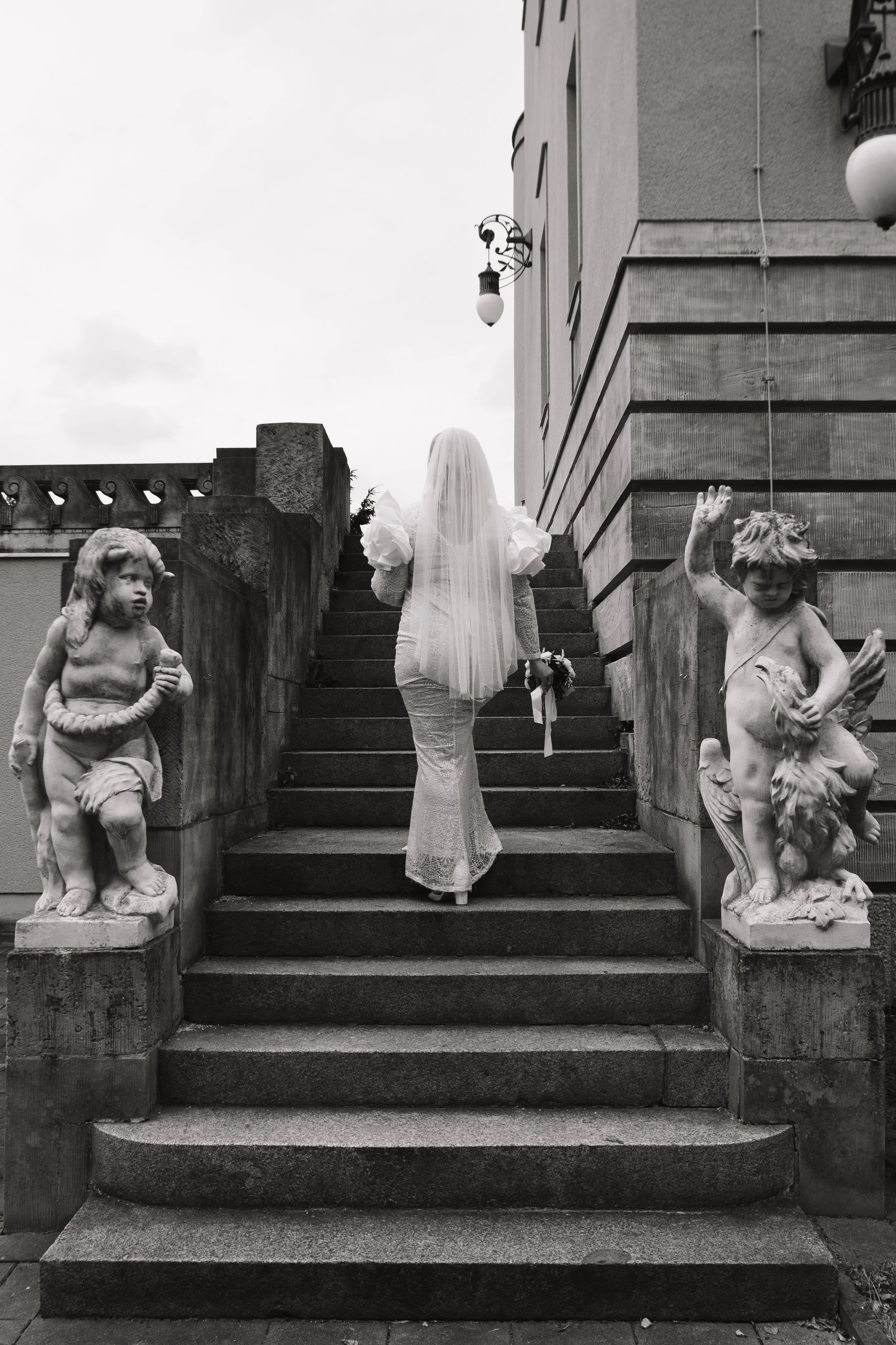 Eine Frau in einem Brautkleid mit Schleier, die die Treppe zu einem Gebäude hinaufsteigt, flankiert von zwei Skulpturen von Kindern auf der linken und rechten Seite.