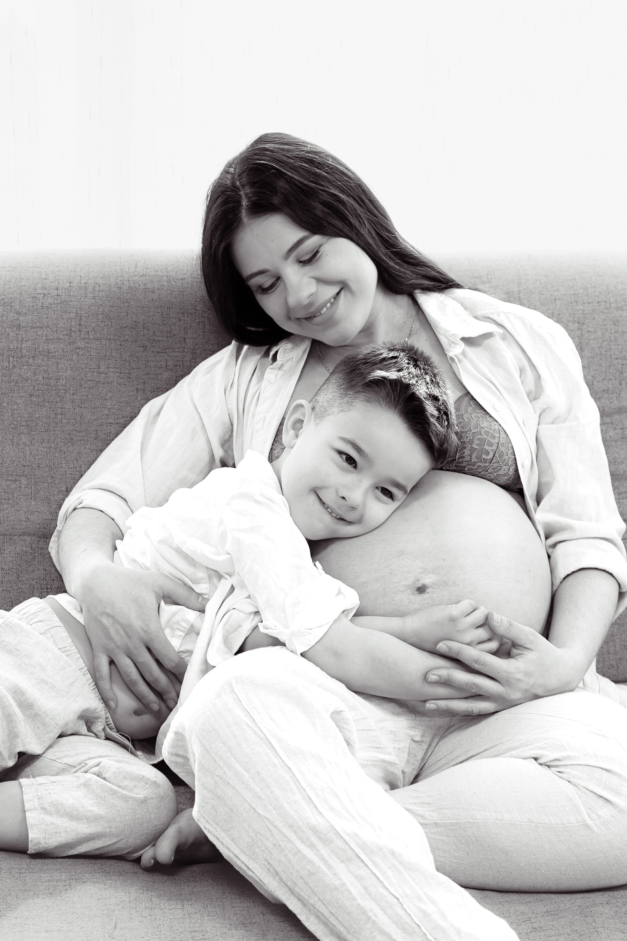 Schwangere Frau sitzt auf einer Couch, lächelt und hält ihrBaby, das auf ihrem Bauch liegt, während ein kleiner Junge kuschelt und lächelt.