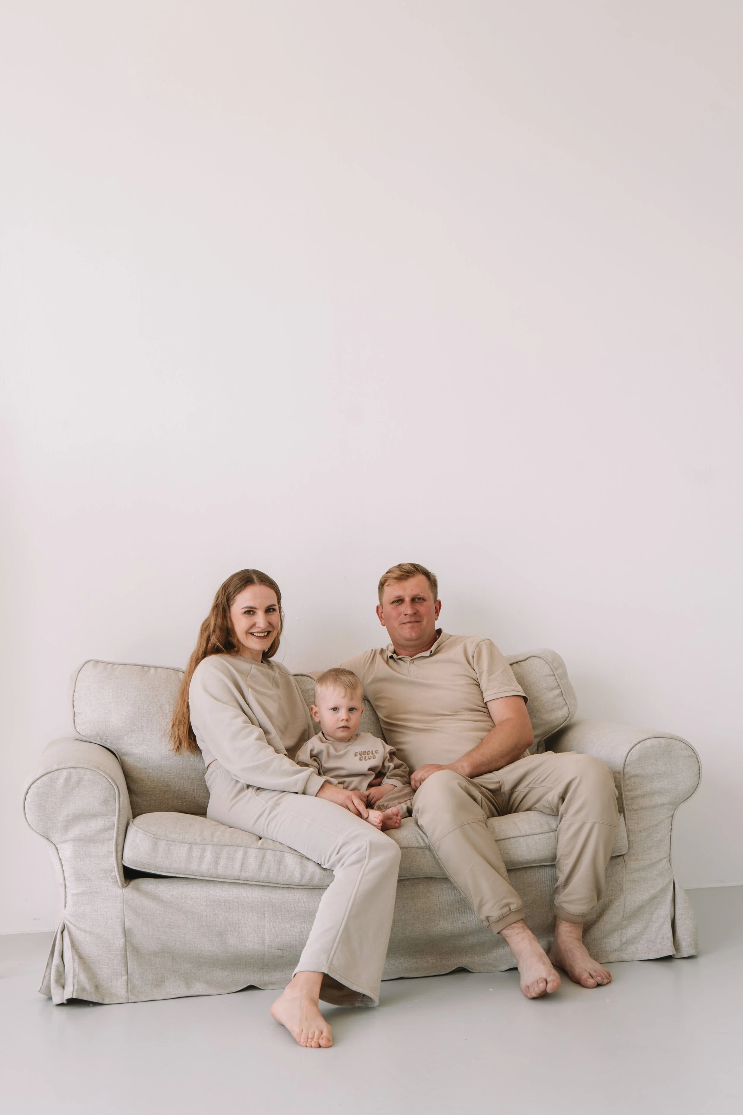 Familie sitzt auf einem beige Couch in einem minimalistischen Raum, alle tragen bequeme, beige Kleidung, Baby sitzt auf dem Schoß der Mutter.
