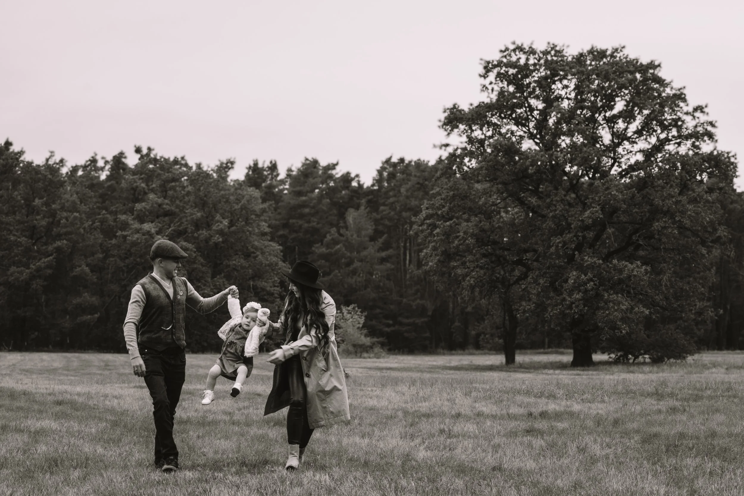 Ein Paar spielt mit einem kleinen Mädchen auf einer Wiese im Freien, umgeben von Bäumen, in Schwarz-Weiß-Fotografie.
