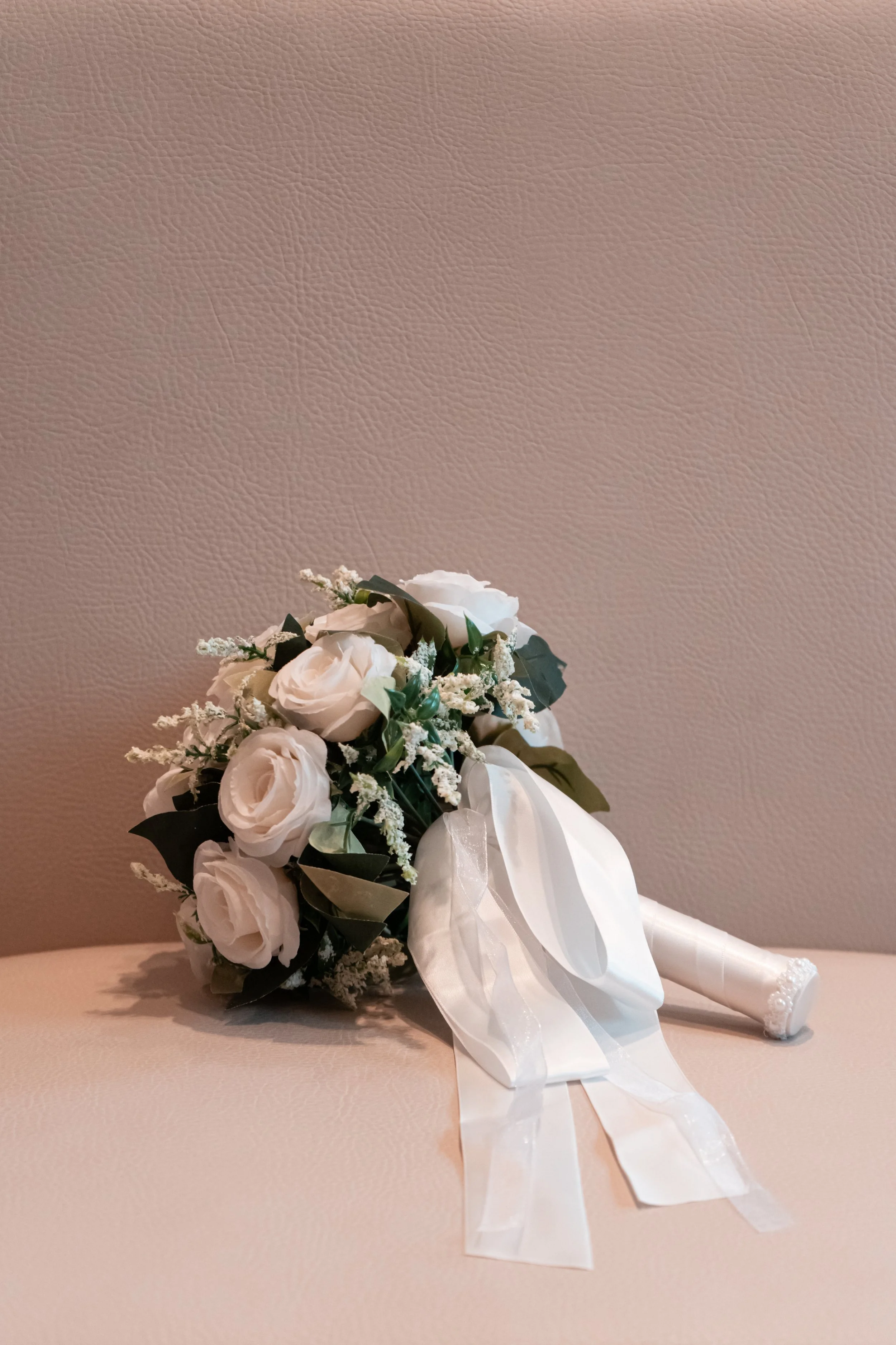 Brautstrauß mit weißen Blumen, Schleife und grünen Blättern auf gepuderter Oberfläche.